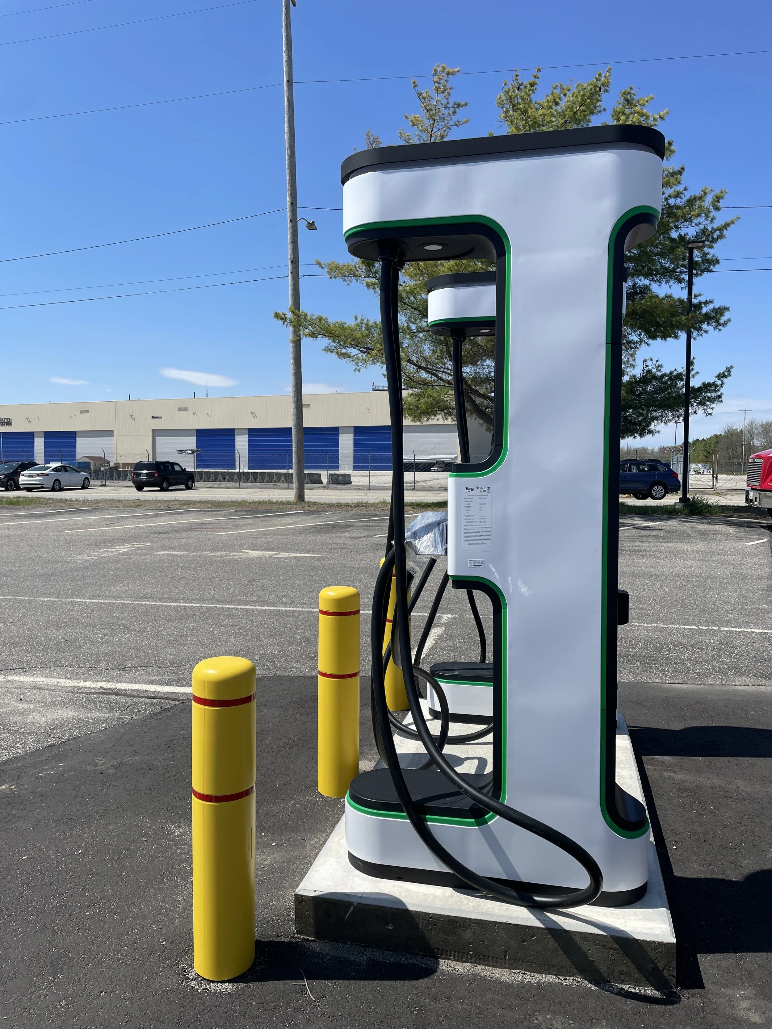 An electric vehicle charging station in a parking lot under a clear blue sky, with yellow safety bollards in the foreground and a building with blue and white exterior in the background.
