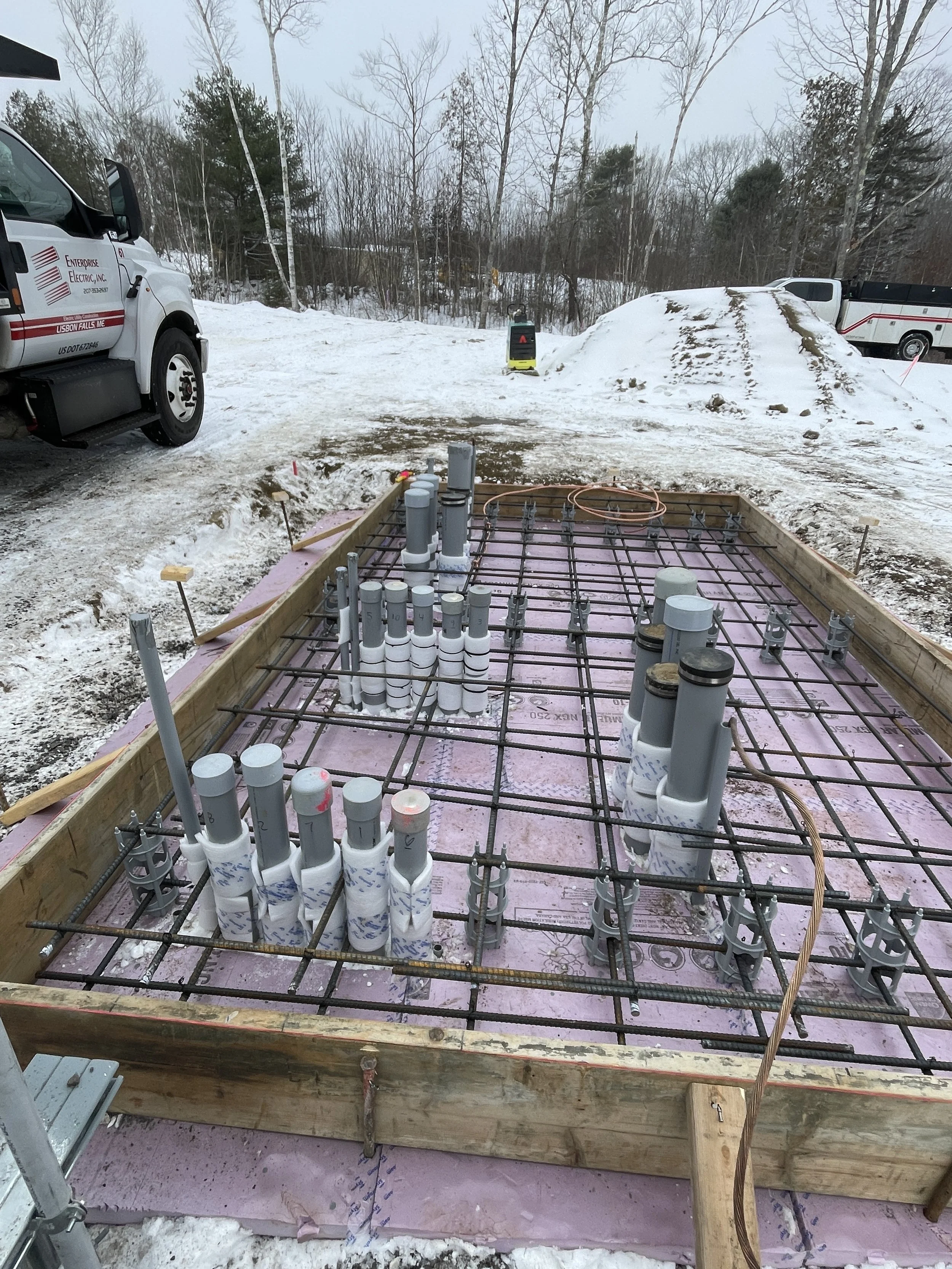Construction site in winter with PVC pipes for plumbing, concrete rebar framework, and snow-covered ground, surrounded by trees, with utility vehicles parked nearby.