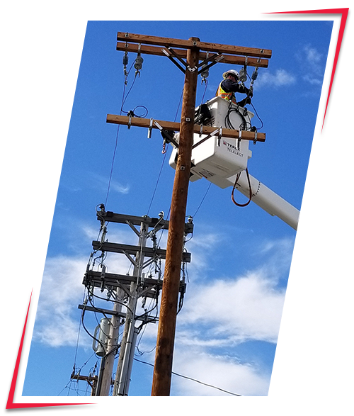 A worker in safety gear working on power lines from a cherry picker bucket elevated by a utility truck, performing maintenance or repairs on a wooden utility pole.