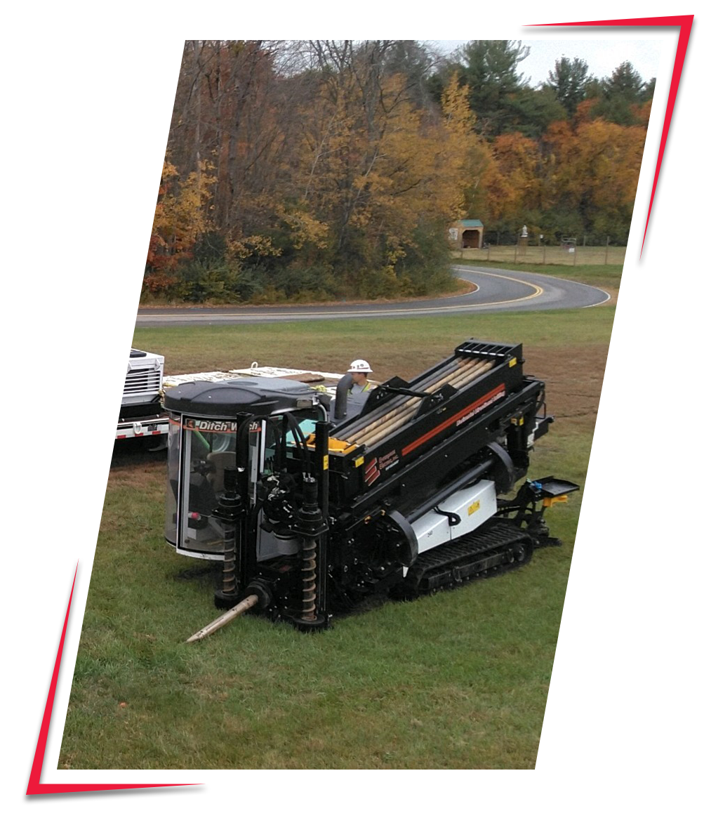 A tracked drilling machine with a hydraulic arm and a long drill bit parked on grass, with two workers nearby wearing helmets.