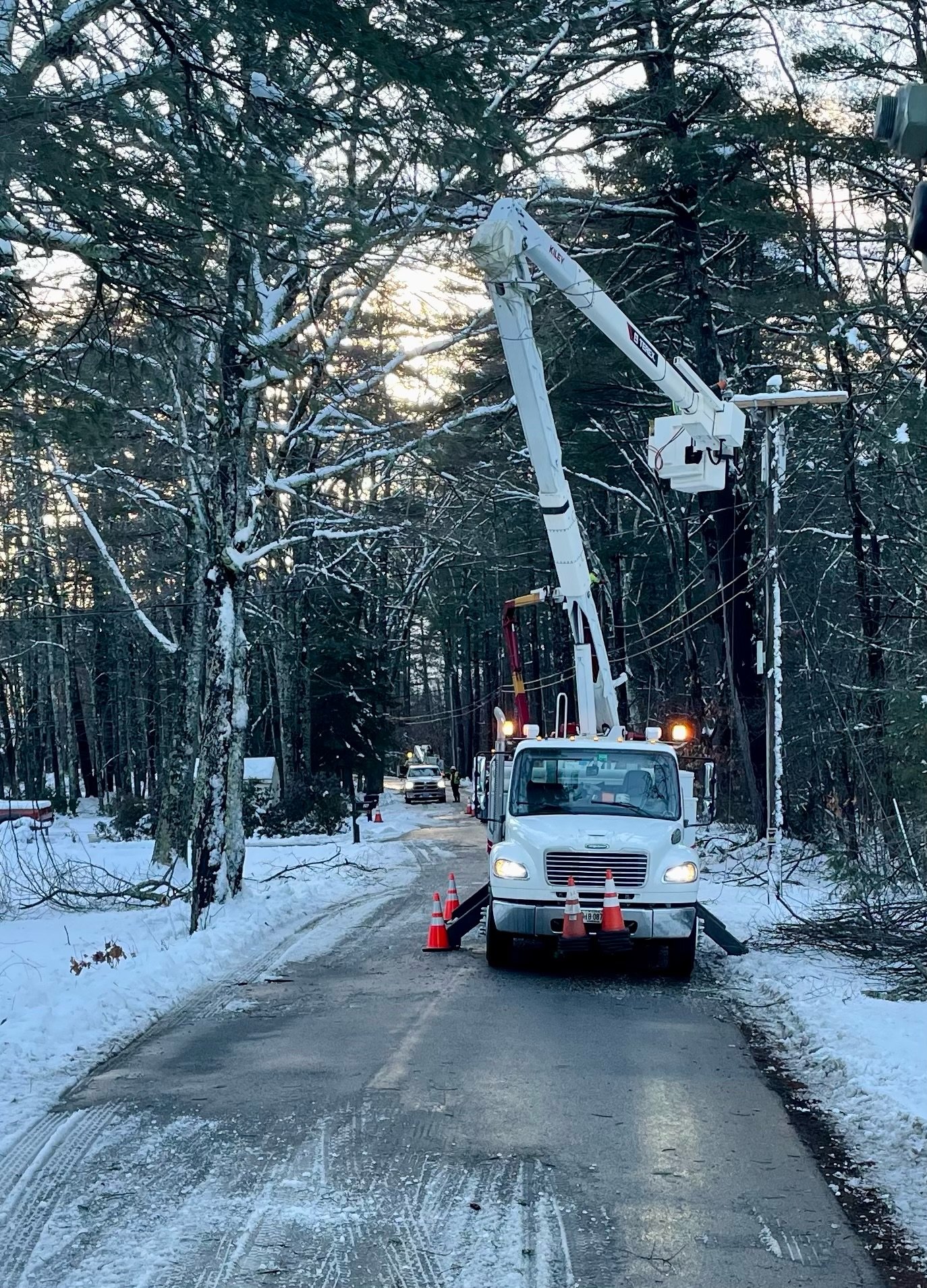 Utility workers in a bucket lift repairing a power line on a snow-covered road surrounded by trees.
