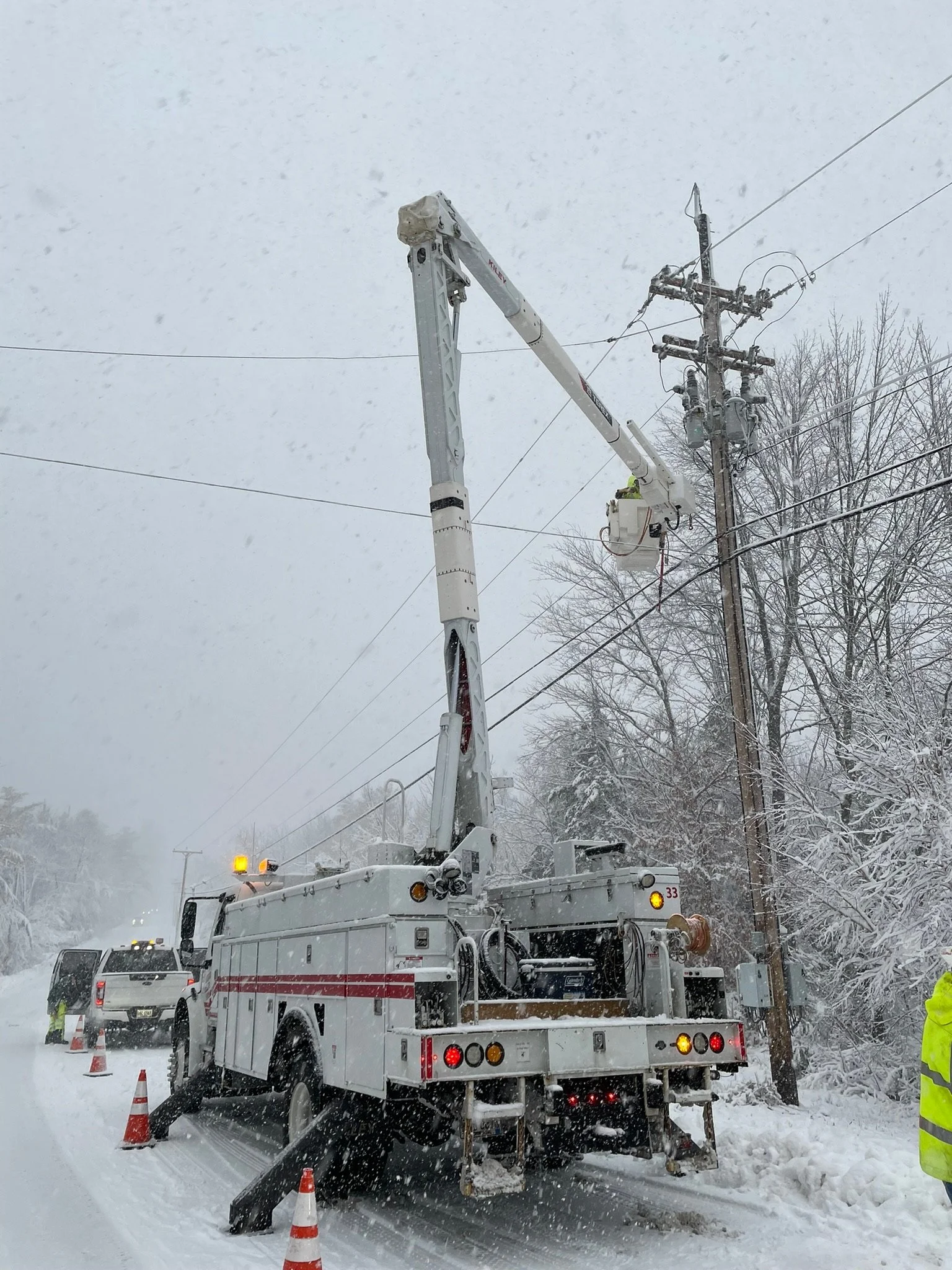 A utility truck with an extended cherry picker working on power lines during a snowy day, with traffic cones placed around for safety.