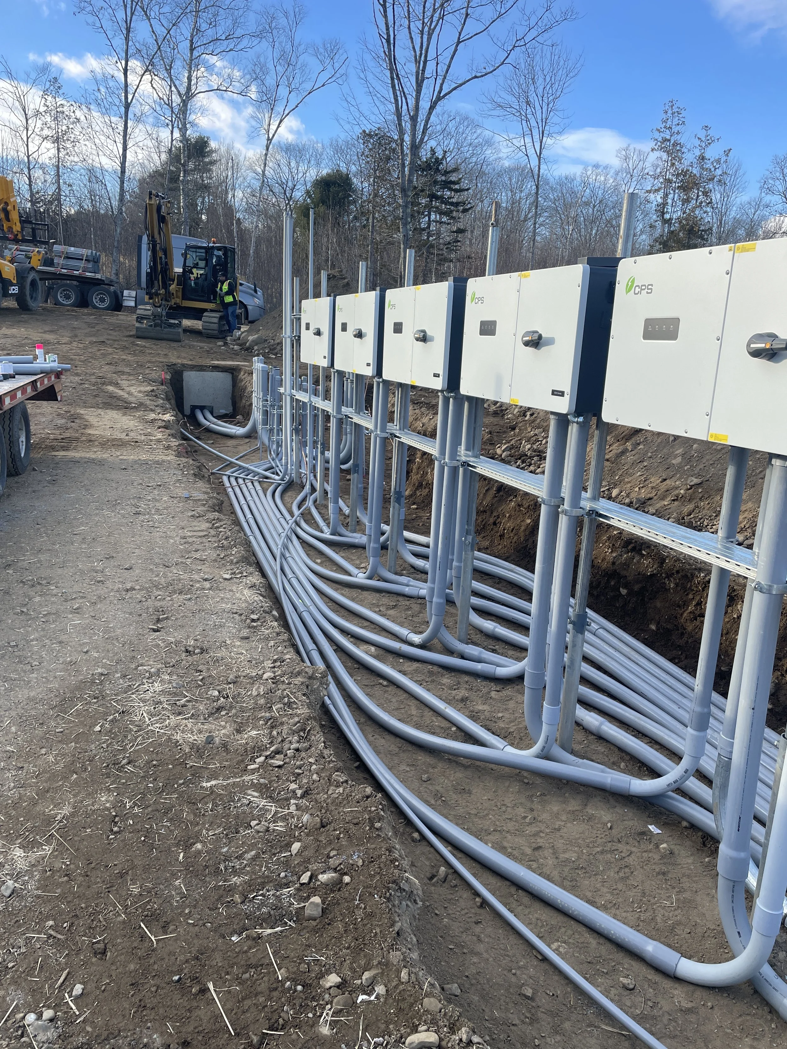 Construction site with electrical boxes, pipes, and machinery, surrounded by dirt and bare trees under a blue sky.