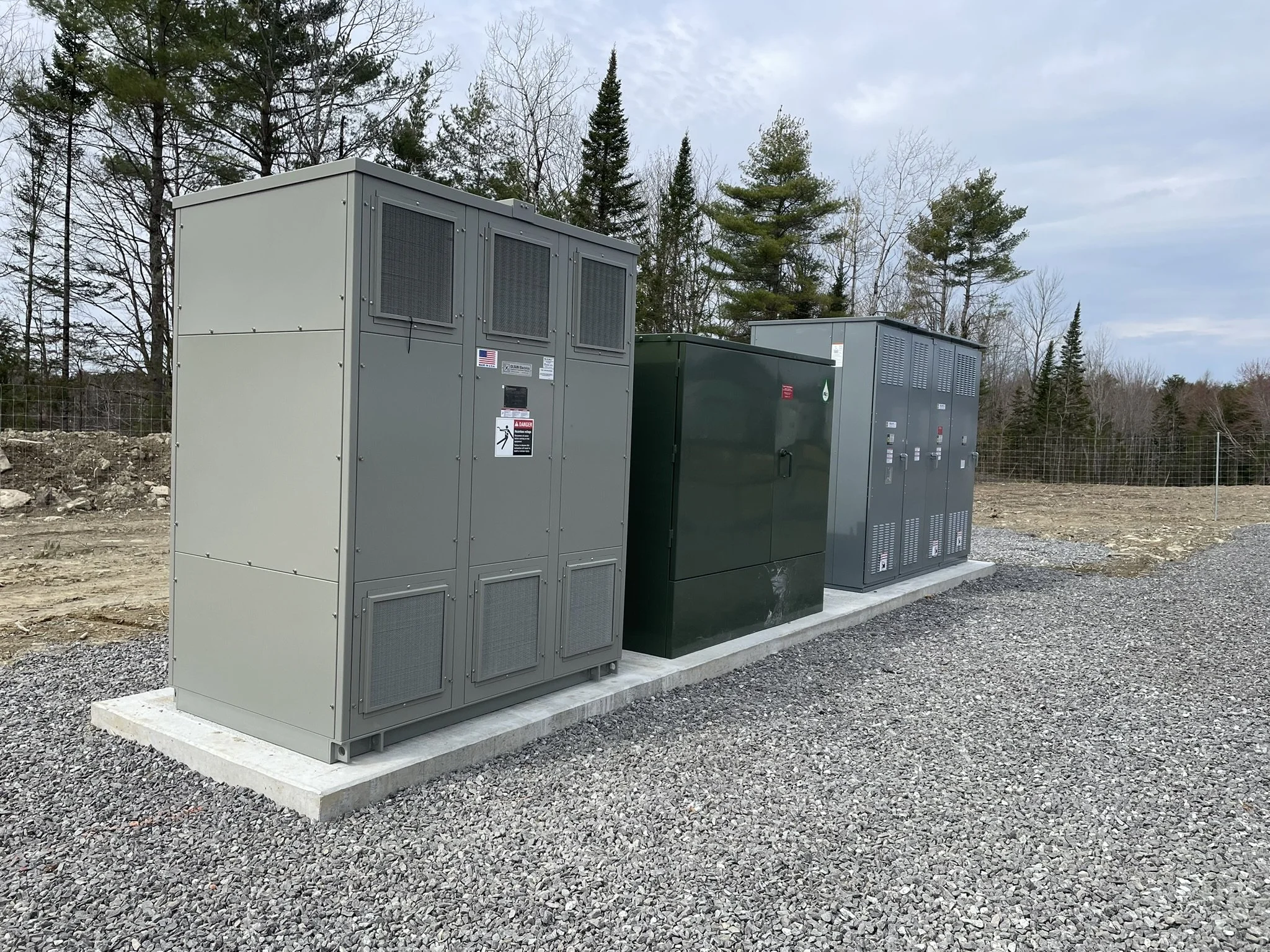 Electrical transformers and a green utility box installed on concrete slab outdoors with trees and cloudy sky in background.