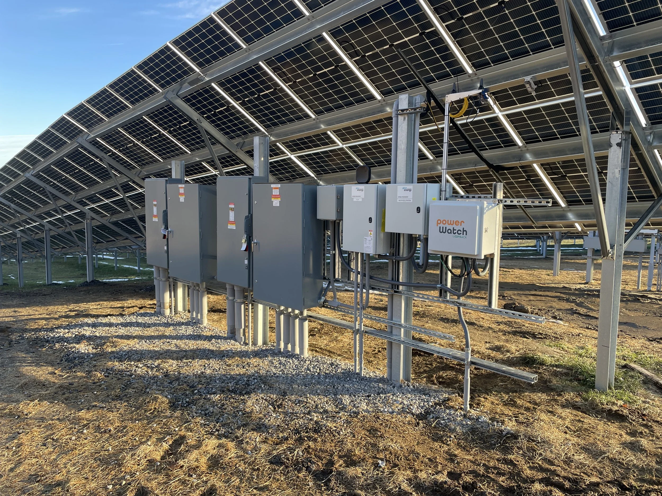 Solar panels installed on a structure, with electrical equipment and wires visible in the foreground, set against a clear blue sky.