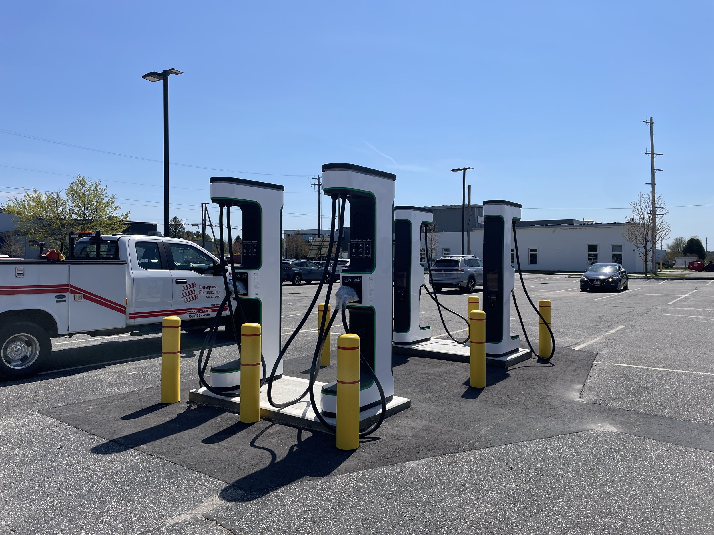 Electric vehicle charging stations in a parking lot with cars, a pickup truck, and a building in the background under a clear blue sky.