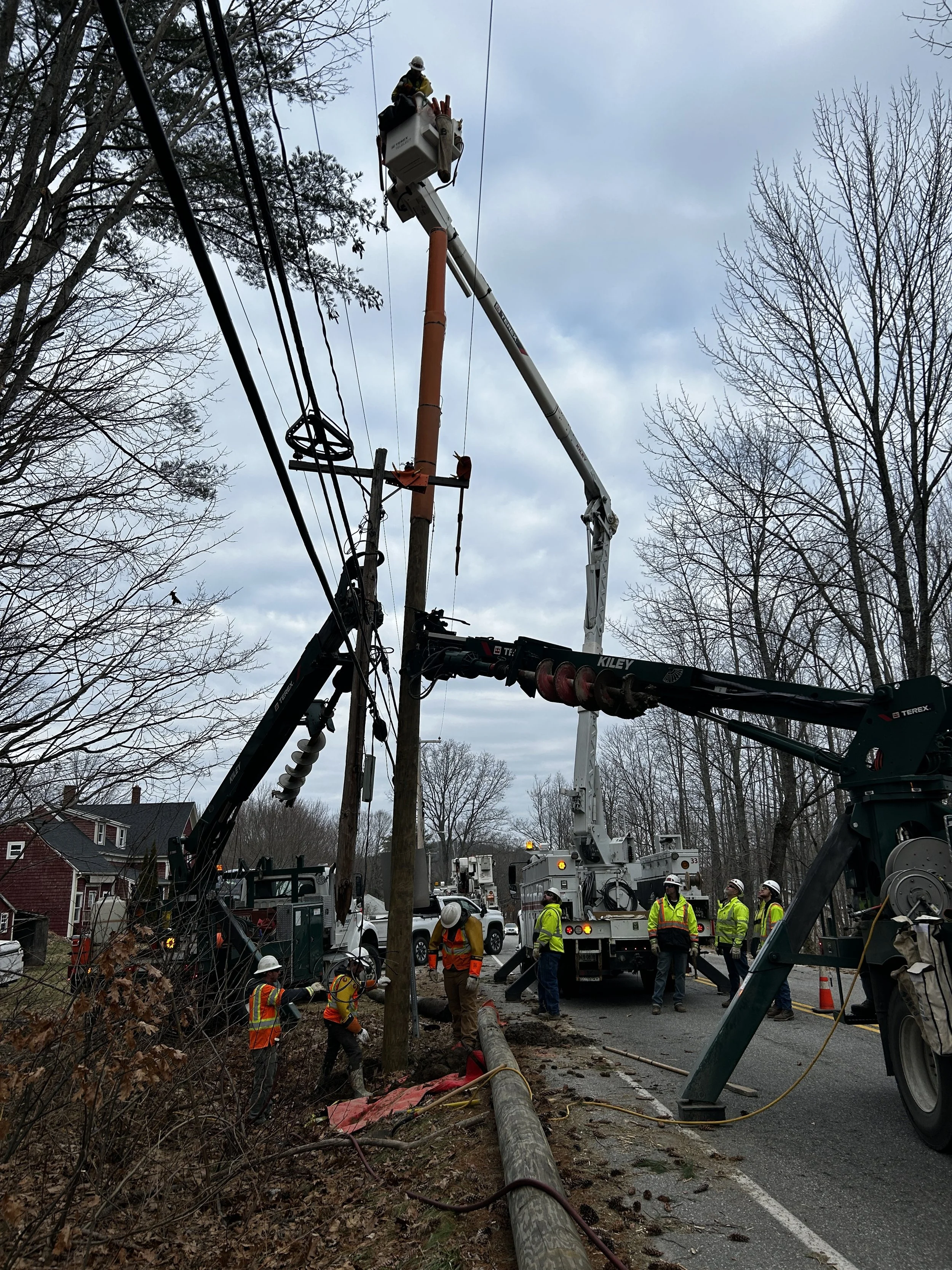 Workers and emergency personnel repair a fallen utility pole with a crane and bucket lift outdoors on a cloudy day, with trees and houses in the background.