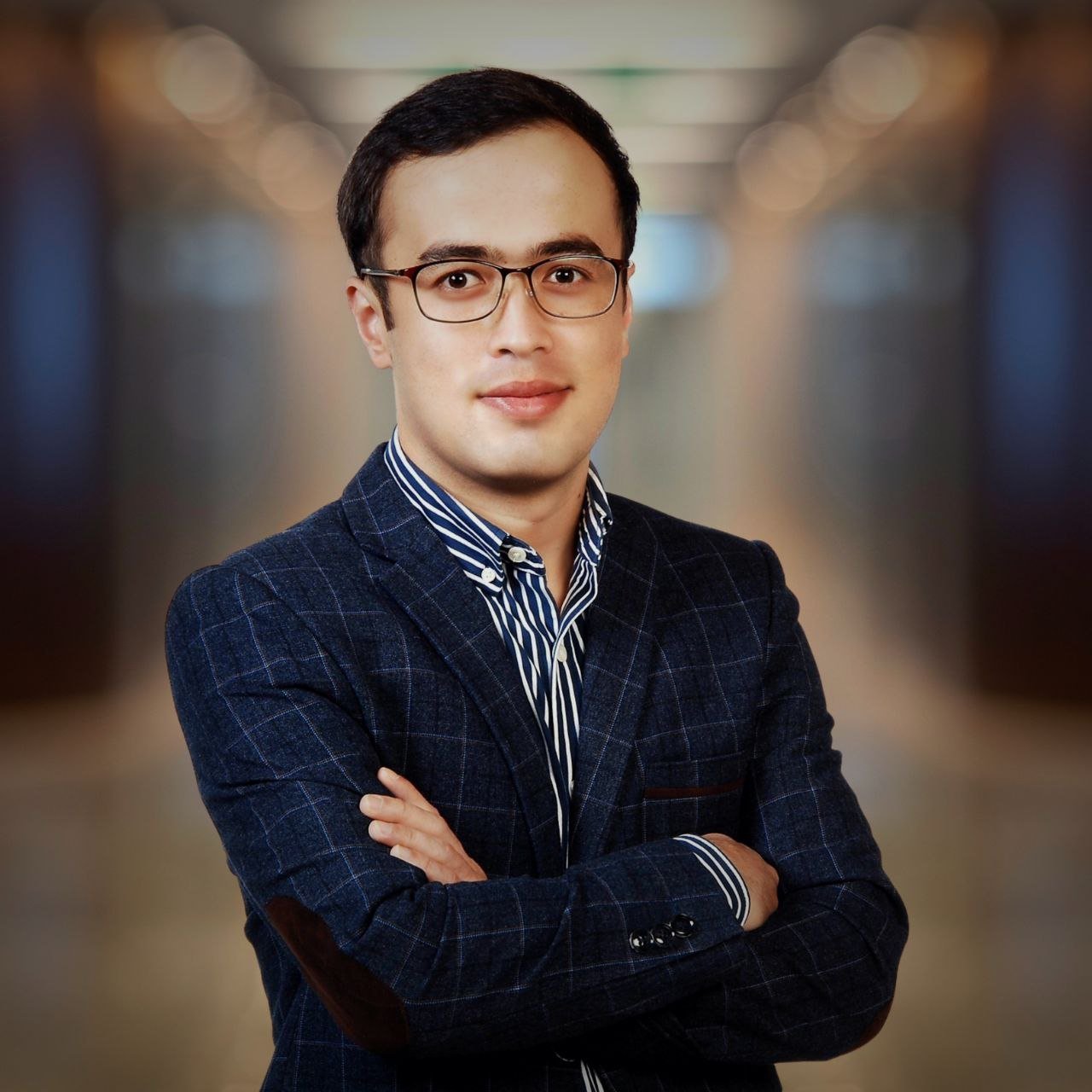 Portrait of a young man with glasses wearing a dark blue plaid blazer and a striped shirt, standing with arms crossed in a blurred indoor setting.