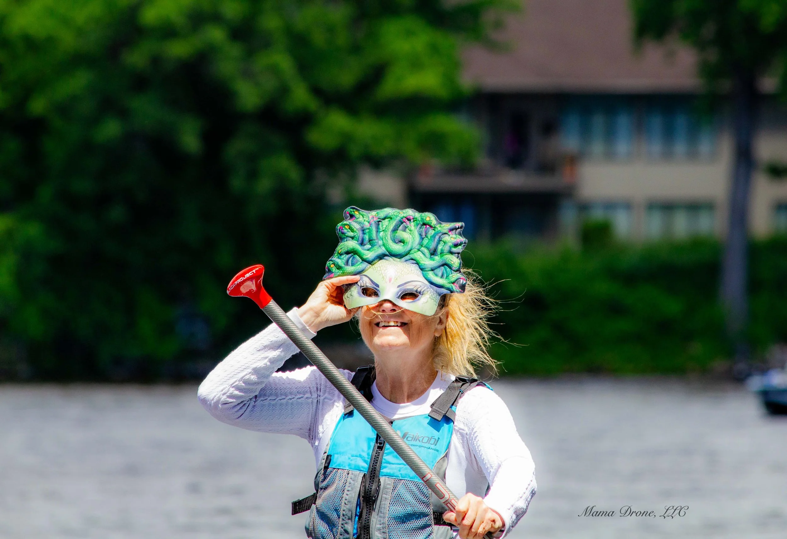 Blonde haired person with masquerade mask and light blue and gray life jacket, holding a red handled canoe ore on open water.