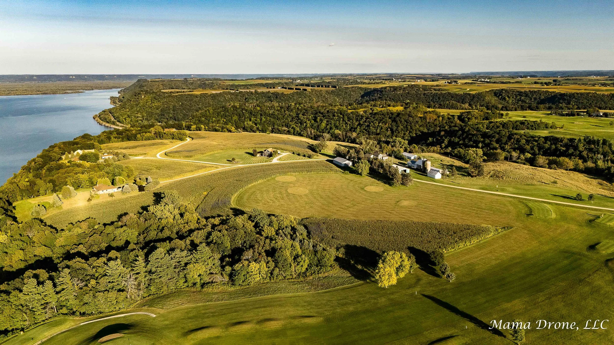 Aerial view of a bluff top golf course, blue open water, green trees and neighboring houses.