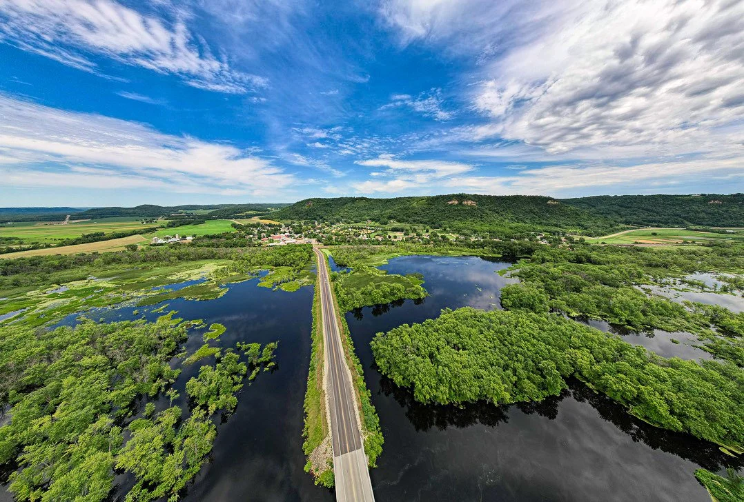 Summer Drone Photography: Wisconsin from the air. Capturing a rural road framed by blue summer skies, a quiet river, and vibrant green marshland.