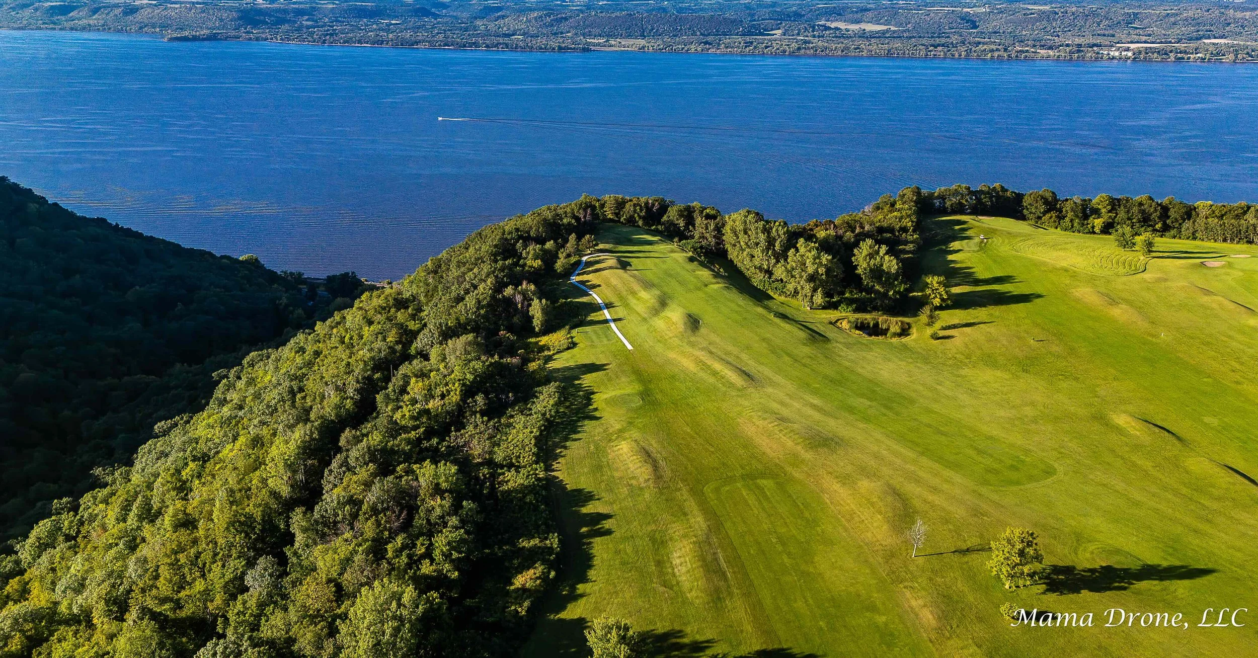 Green trees and grass bluff top golf course view of vibrant royal blue river and land in the foreground.