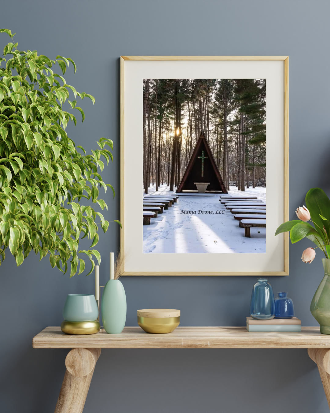 Room wall with green tree, natural wood sofa table with white candles, gold, green and variety of blue vases and pink flowers sitting on it.  A large, natural wood framed photo of a snowy, A-frame chapel in the woods during sunset.