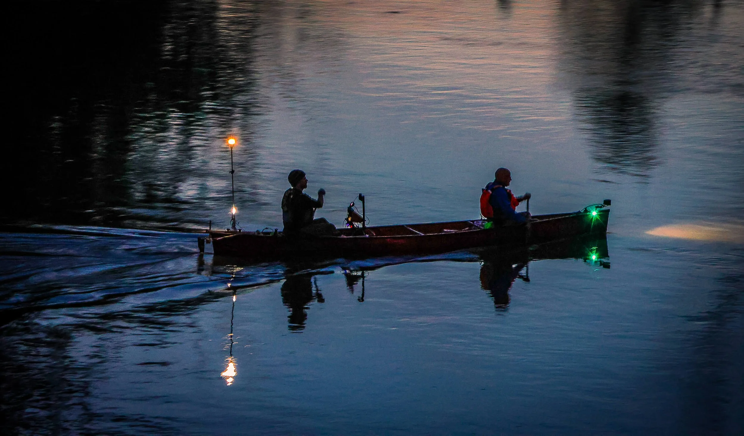 2 people paddling a canoe under lamps on a evening river.