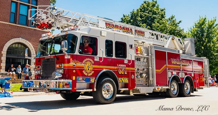Wabasha Fire Department red and white ladder truck with man in red shirt driving it on a street with people watching.