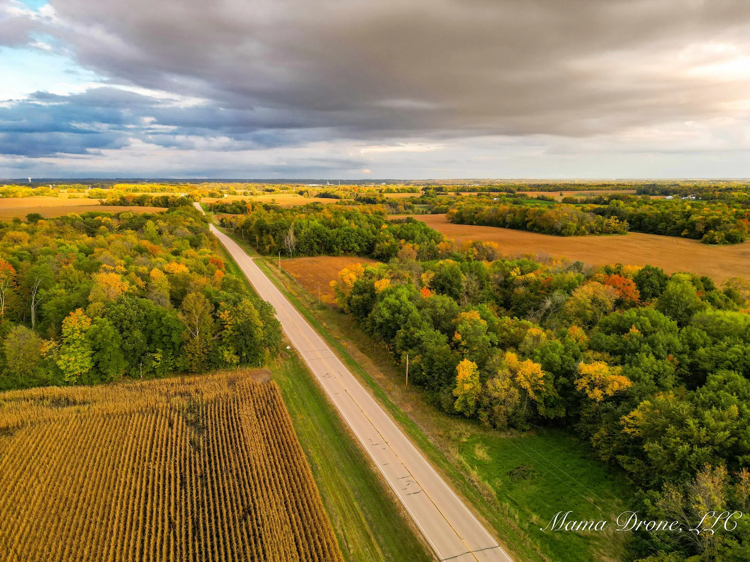 Aerial view of rural paved road intersecting golden farmland and trees of green, yellow and orange.  A gray clouded sky above.