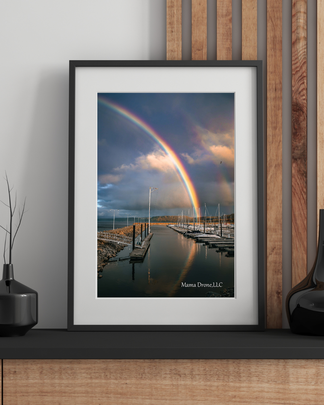 Black framed enlarged photography of a rainbow over a marina, sitting on a wooden ledge with black vases.