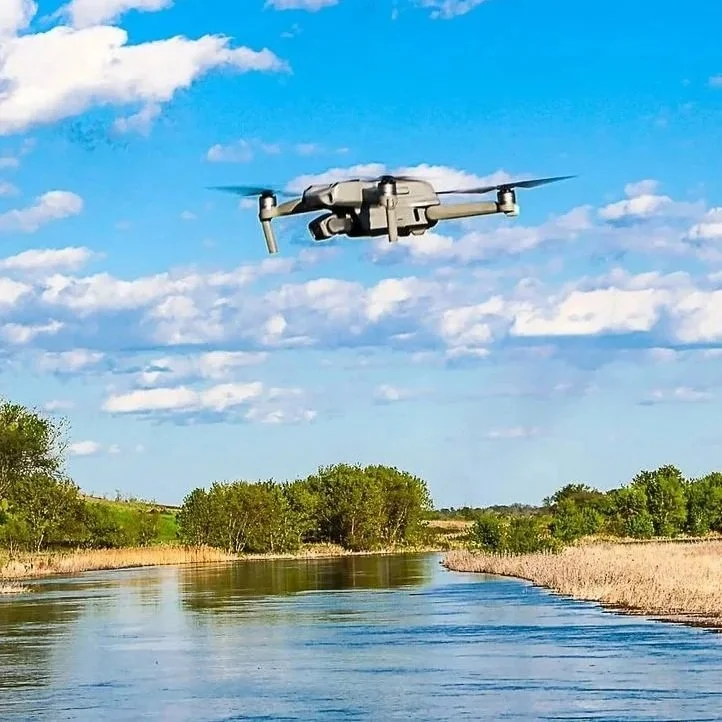 Drone flying over a blue river with tan shorelines, green trees and a cloudy blue sky.
