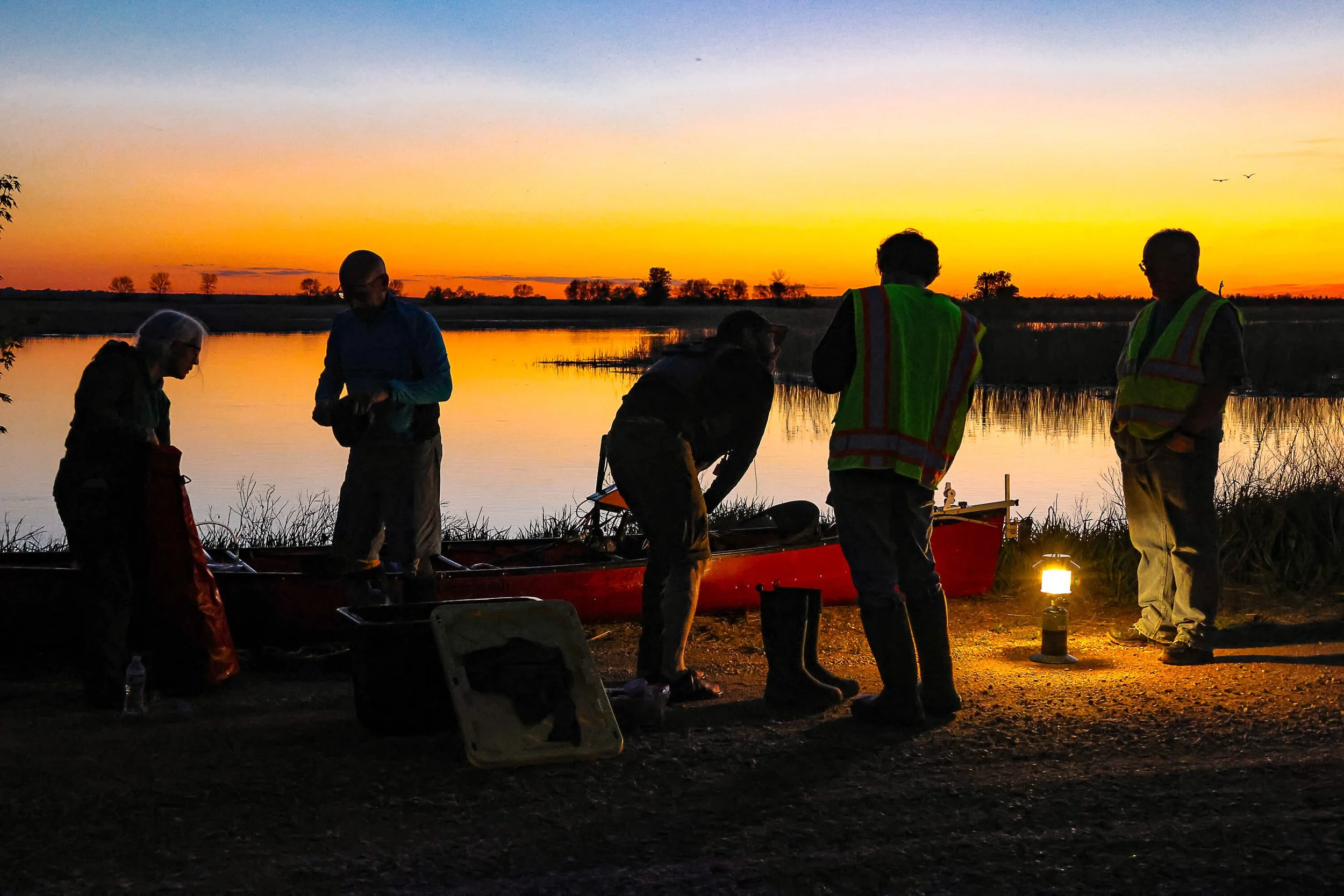 3 men standing near red canoe on a beach with sunset over the water.
