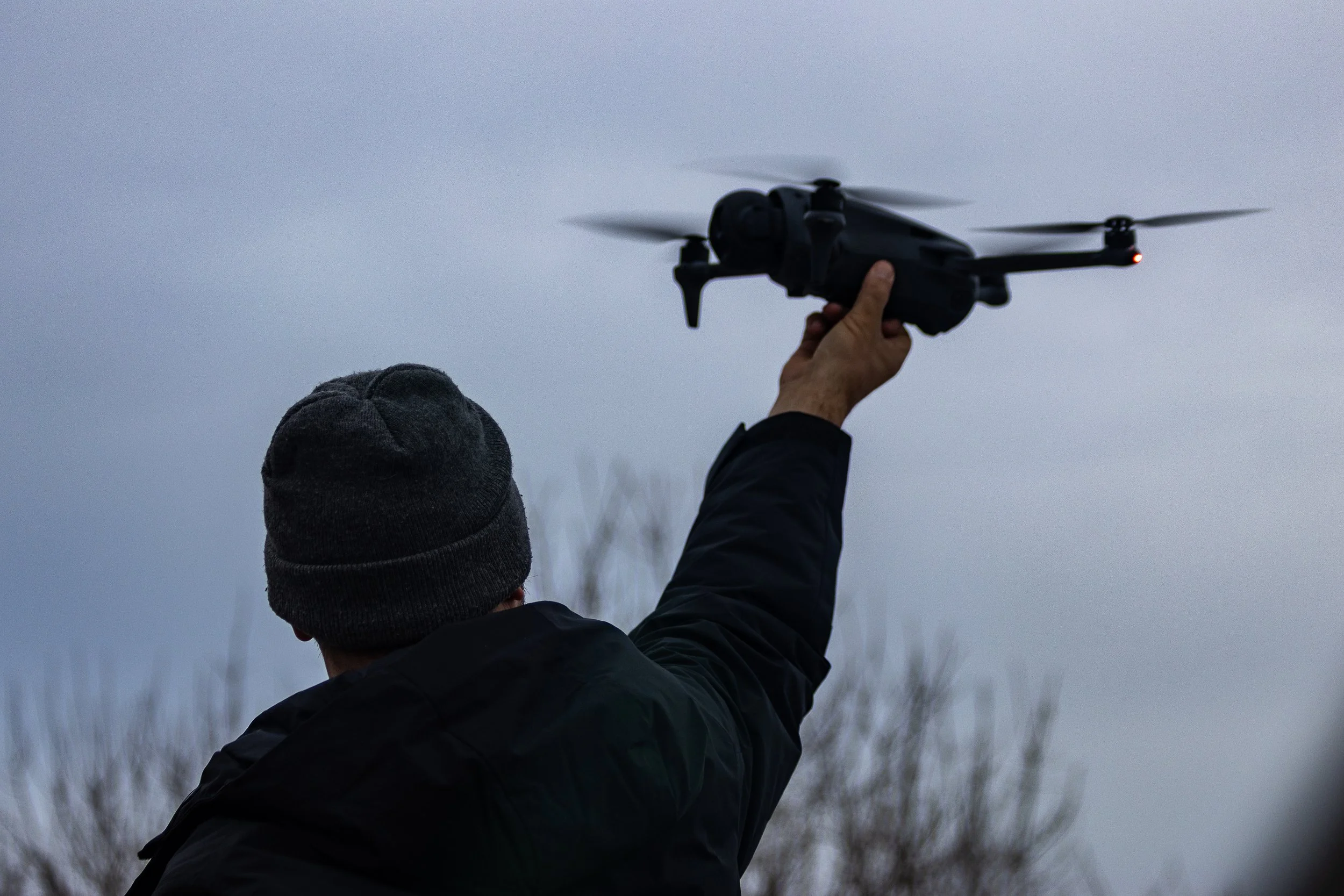 Man with black coat and stocking cap reaching up to hold a black drone.