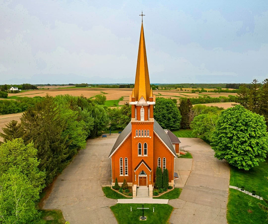 Rural burnt orange colored church with steeple, surrounded by pavement and lush green trees and farmland; under a clear blue sky.