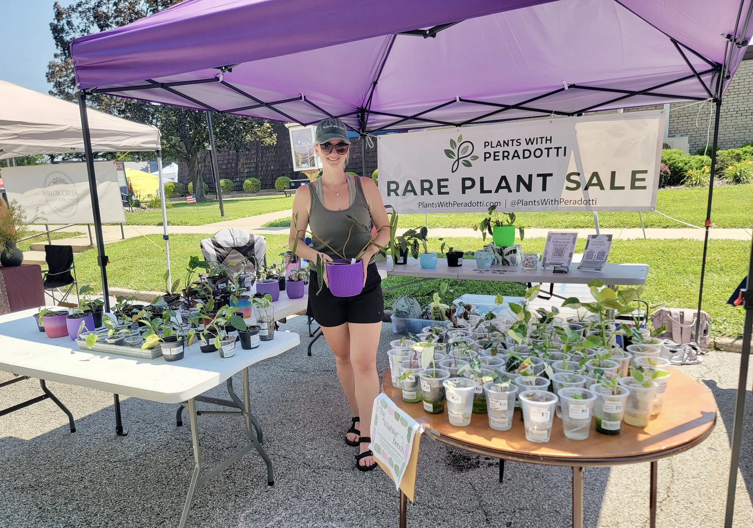 A woman at a plant sale booth, holding a small potted plant, is smiling and wearing sunglasses and a green tank top. The booth has a sign that reads 'RARE PLANT SALE' and displays various potted plants on tables outside.