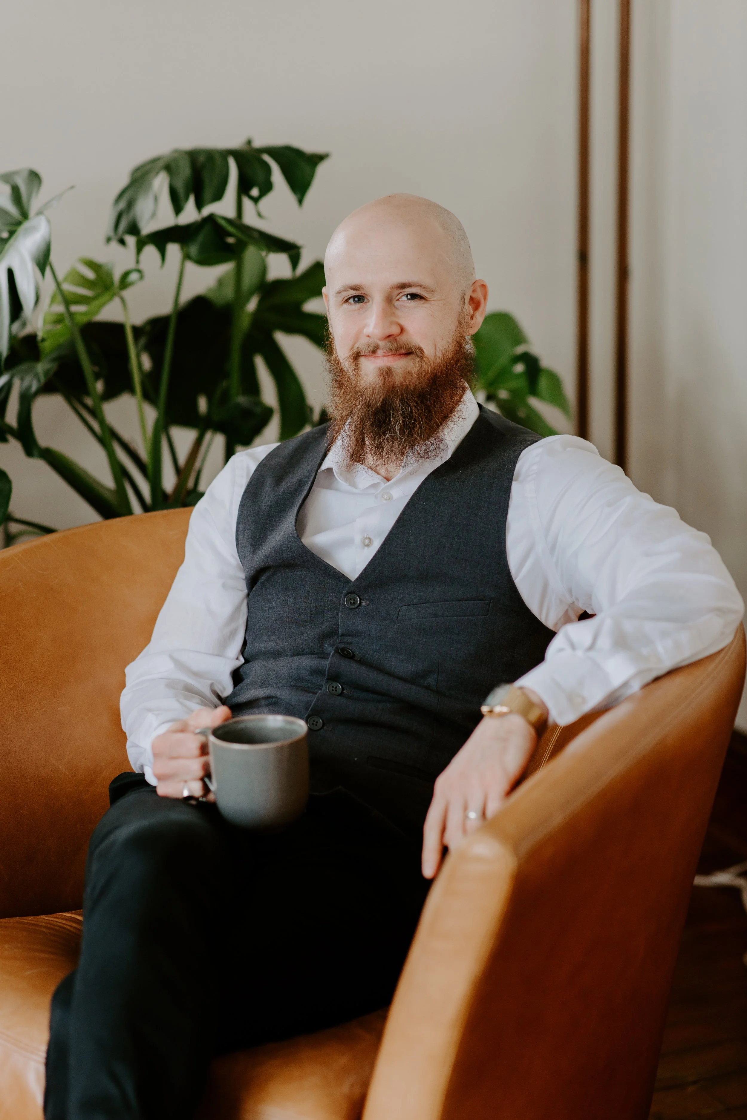 A man with a bald head and a full red beard sitting on a tan leather armchair, holding a gray mug, wearing a white shirt and a dark gray vest, with large leafy green houseplants and wooden frames in the background.