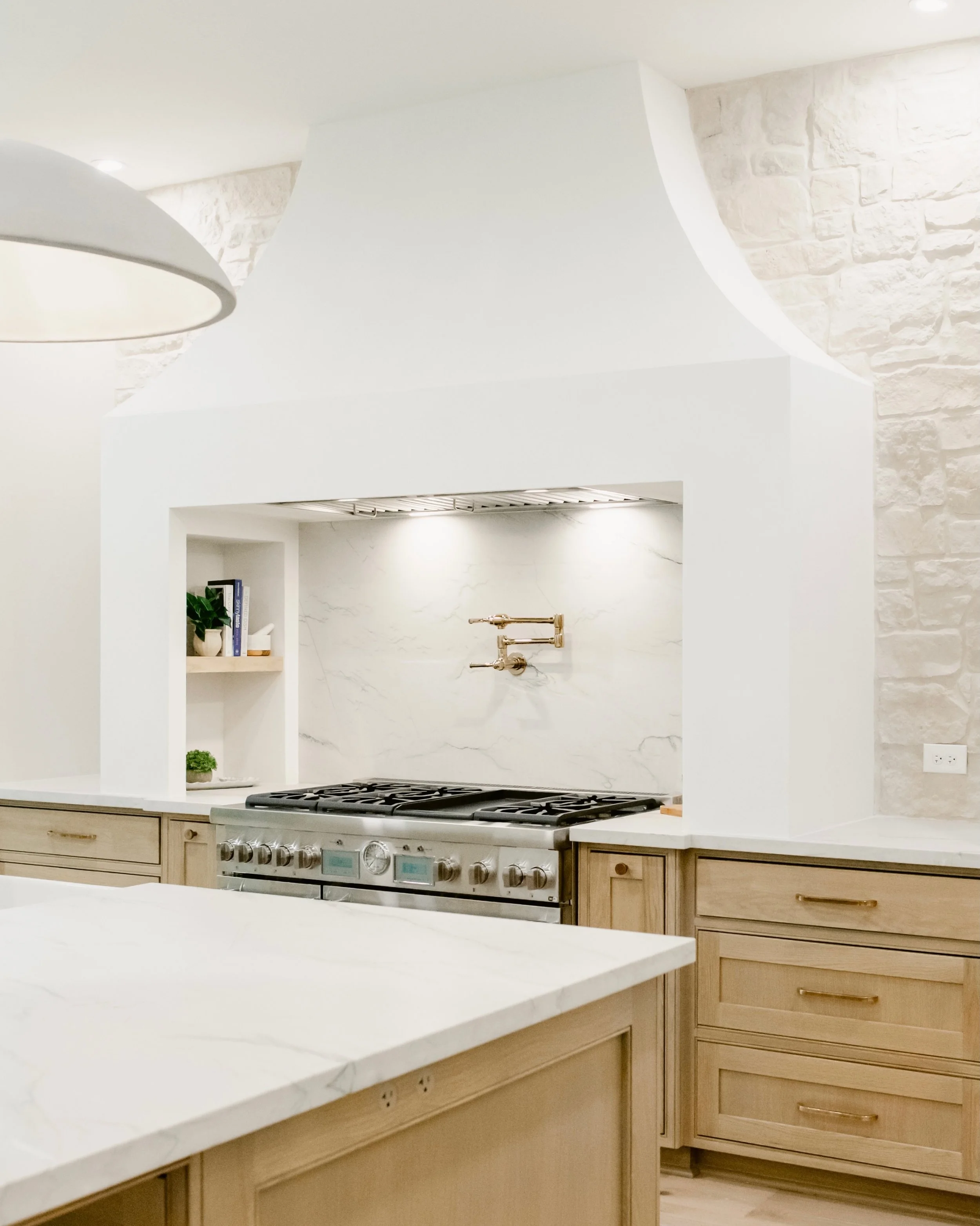 How do you keep an all-white kitchen from feeling cold?

You bring in warmth wherever you can. In this Woodlands kitchen it&rsquo;s white oak cabinets and island, a limestone wall with real texture, light wide-plank floors, and brass pulls to finish 