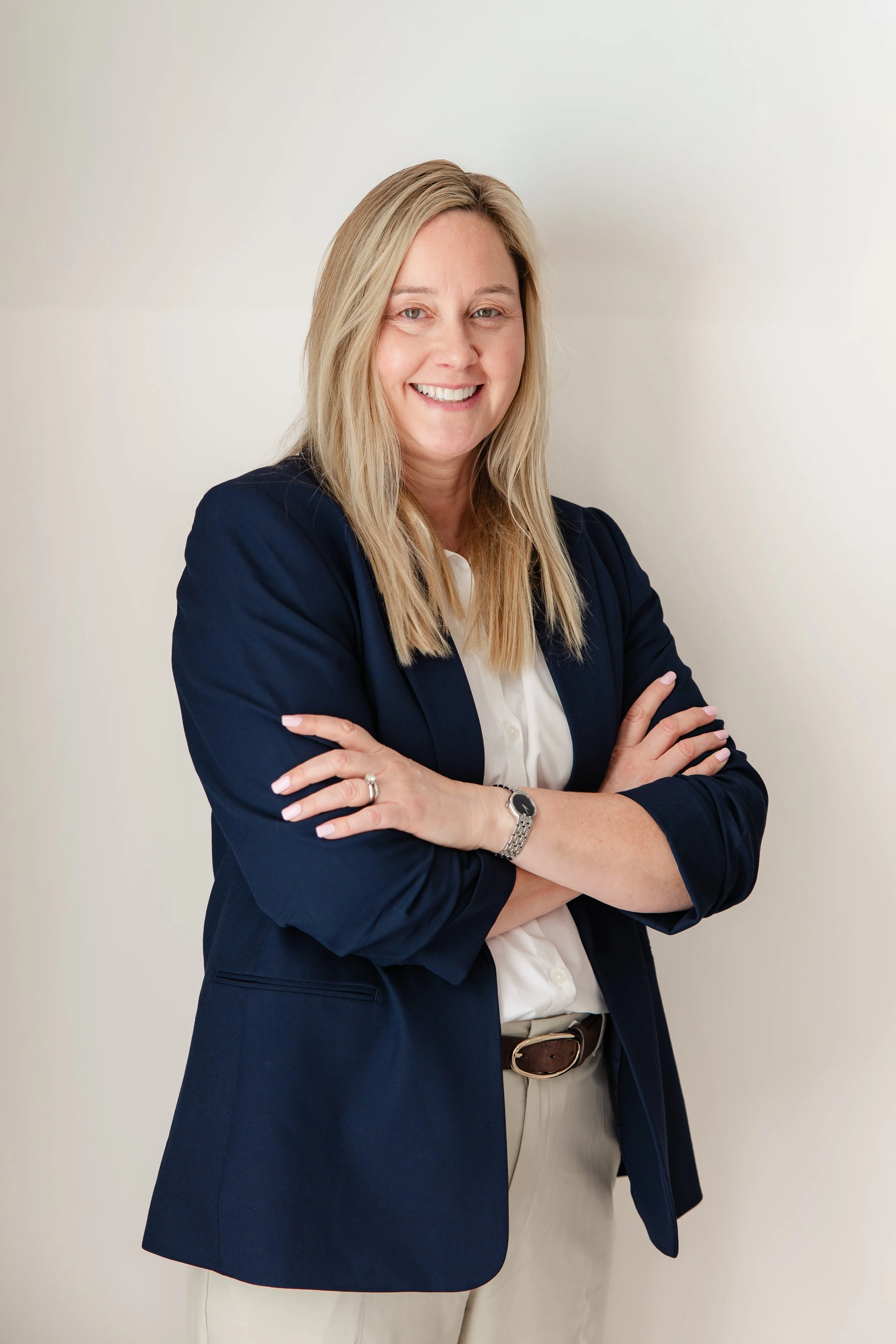 A woman with blonde hair wearing a navy blazer and light-colored pants, smiling with arms crossed, standing against a plain white wall.