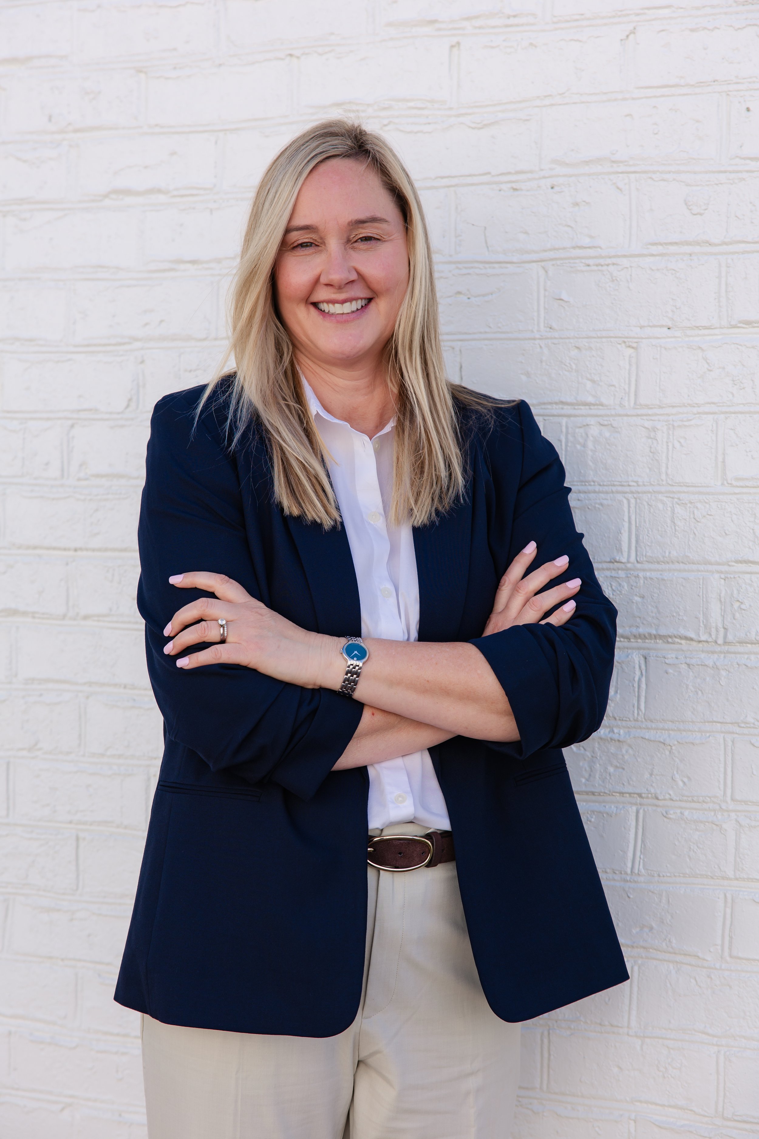 A woman with blonde hair smiling, dressed in a navy blazer and white shirt, standing with arms crossed against a white brick wall.
