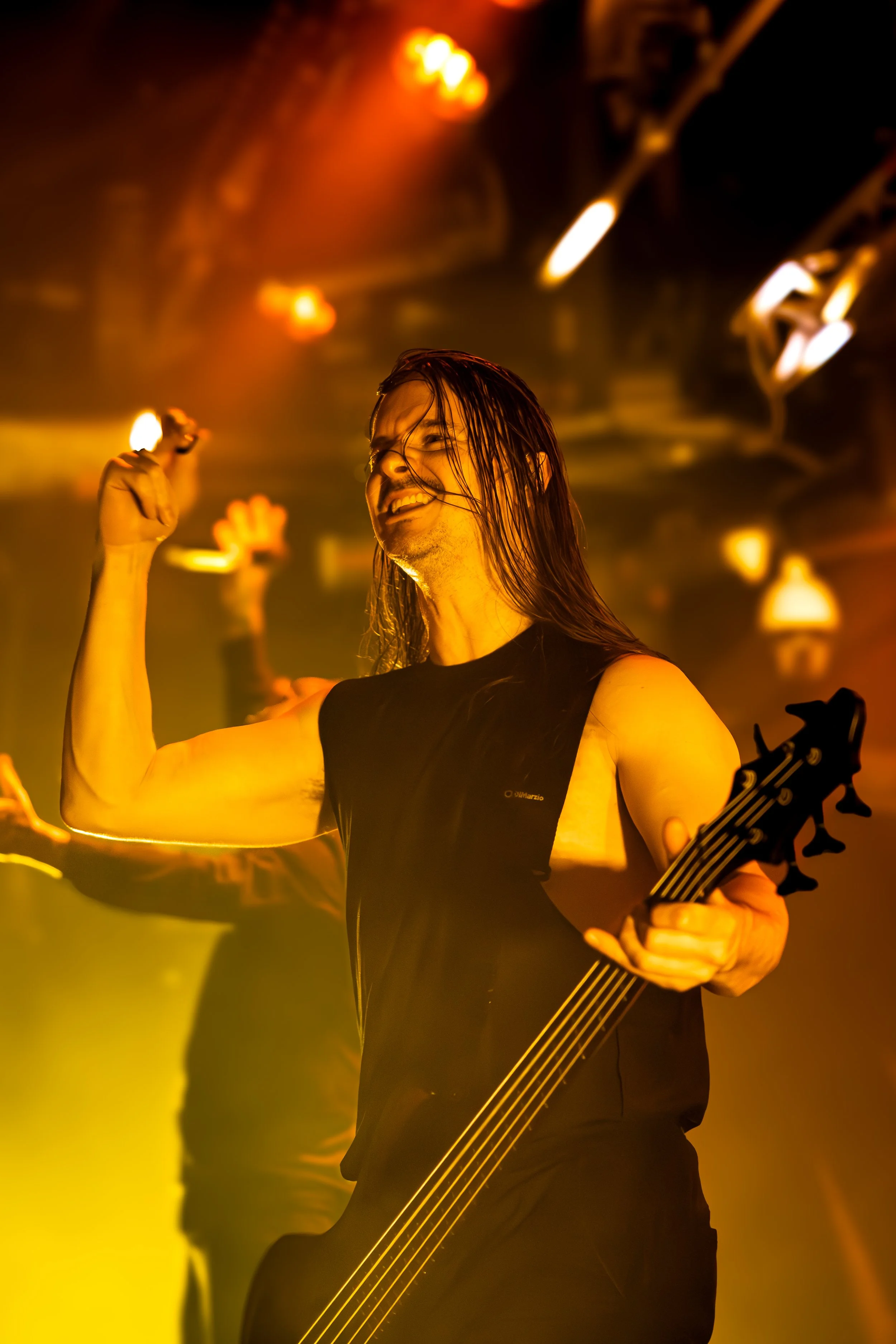 A man with long hair and a beard smiling while playing an electric guitar in a club with colorful stage lighting.