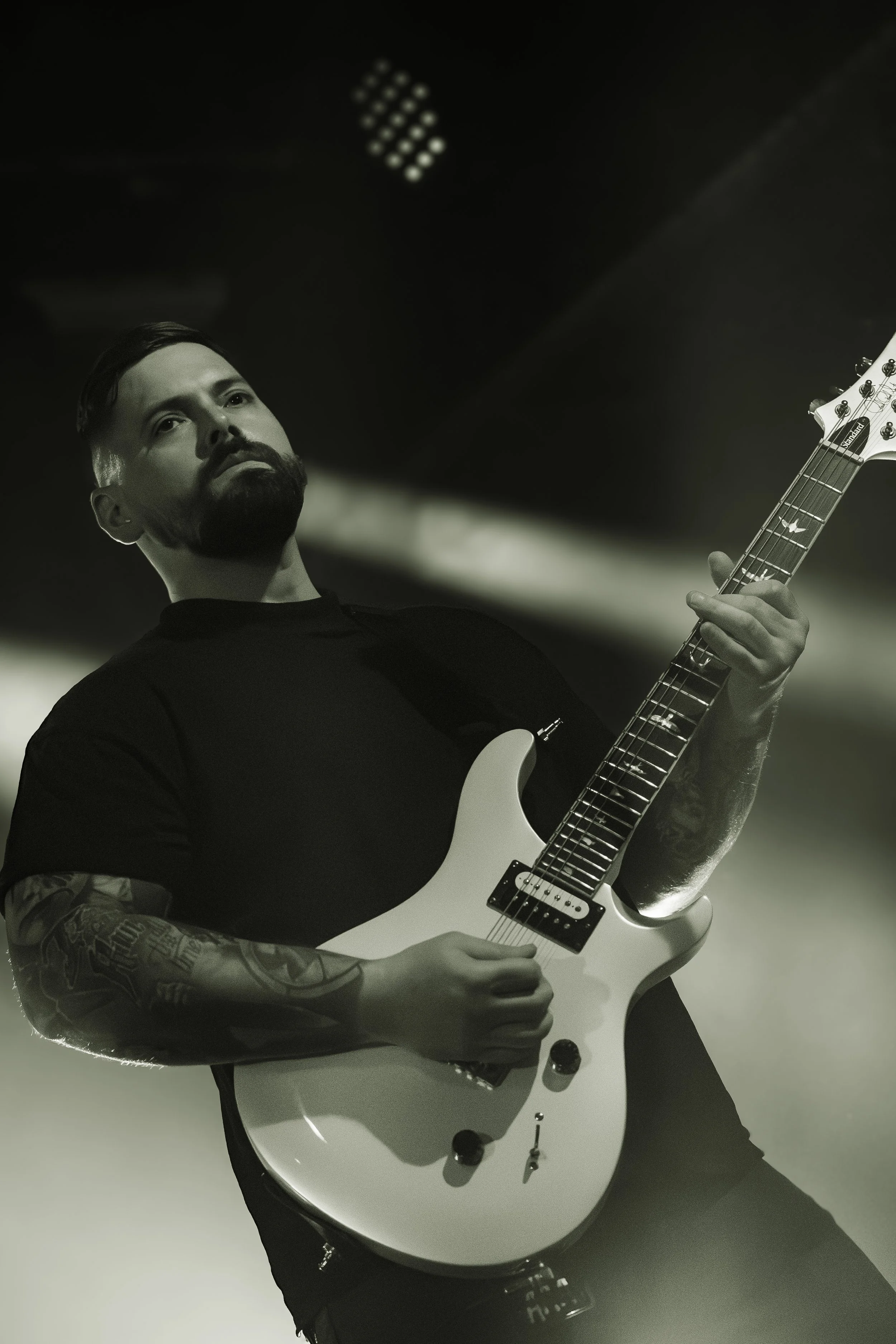 A man with a beard and tattoos on his arm playing an electric guitar. He is looking up in a black and white photo against a blurred background.