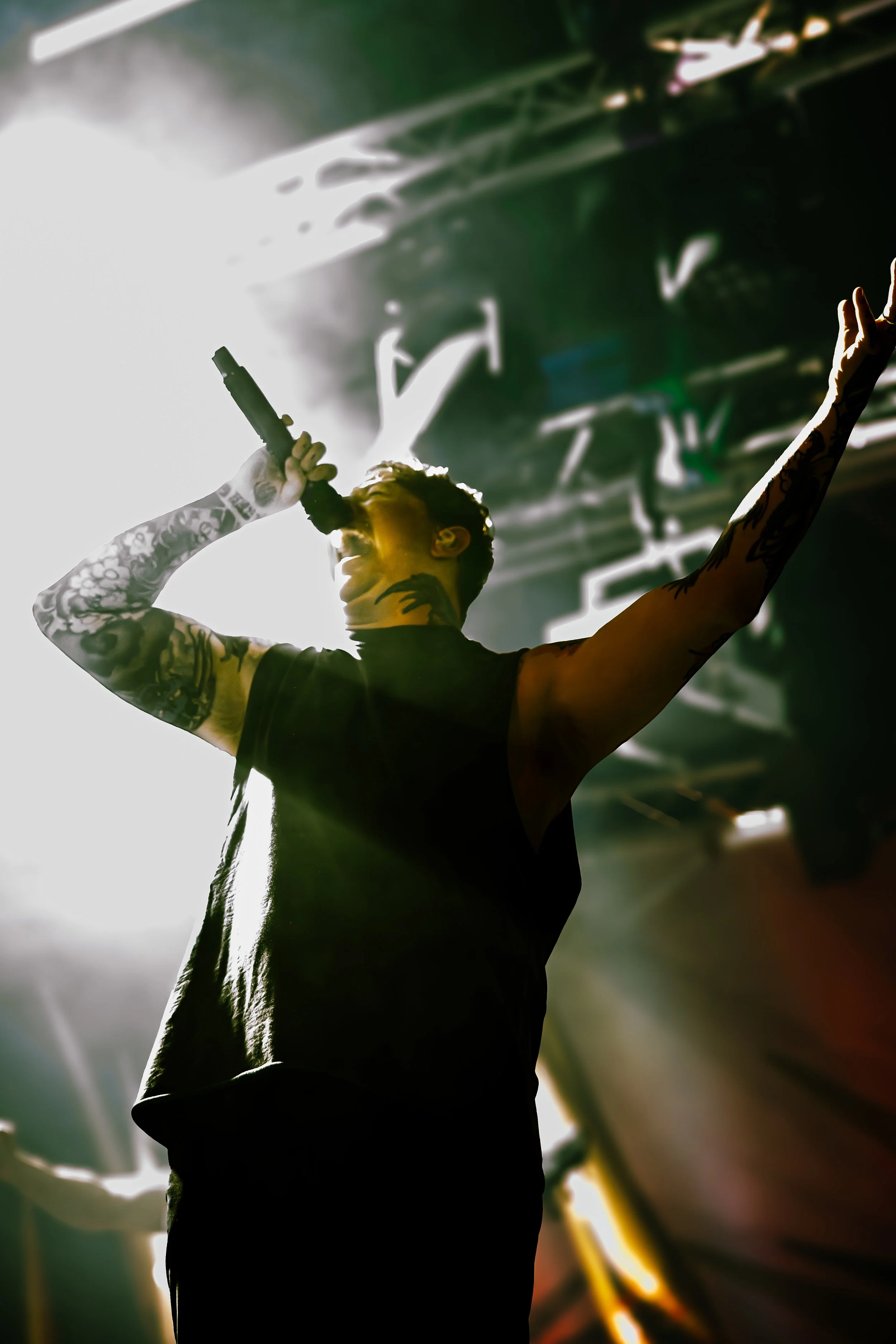 A person with tattoos holding a microphone and singing or speaking on stage with bright backlighting and large green plant leaves in the background.