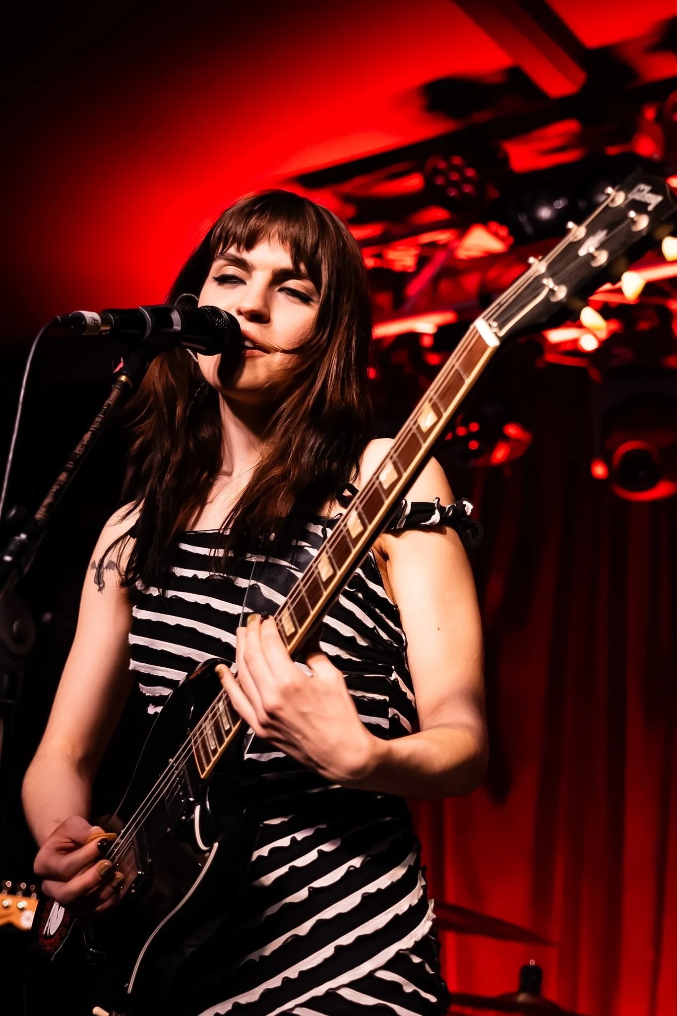 A young woman with dark brown hair and bangs plays an electric guitar on stage, singing into a microphone, with red lighting and stage equipment in the background.