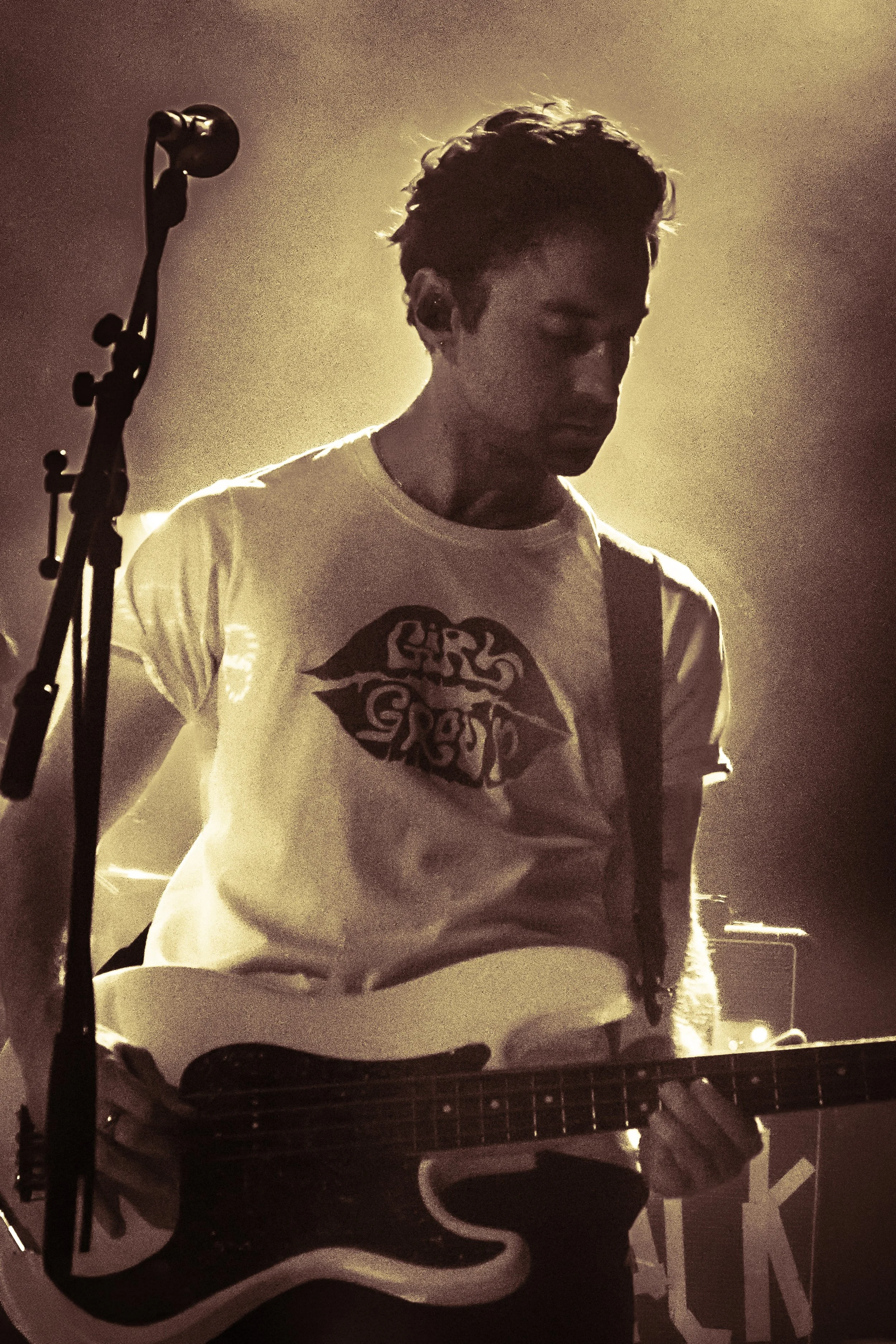 A young man with dark, messy hair playing an electric guitar. He is wearing a white t-shirt with a logo or design on it. The scene appears to be backstage or on stage with warm lighting creating a silhouette effect.