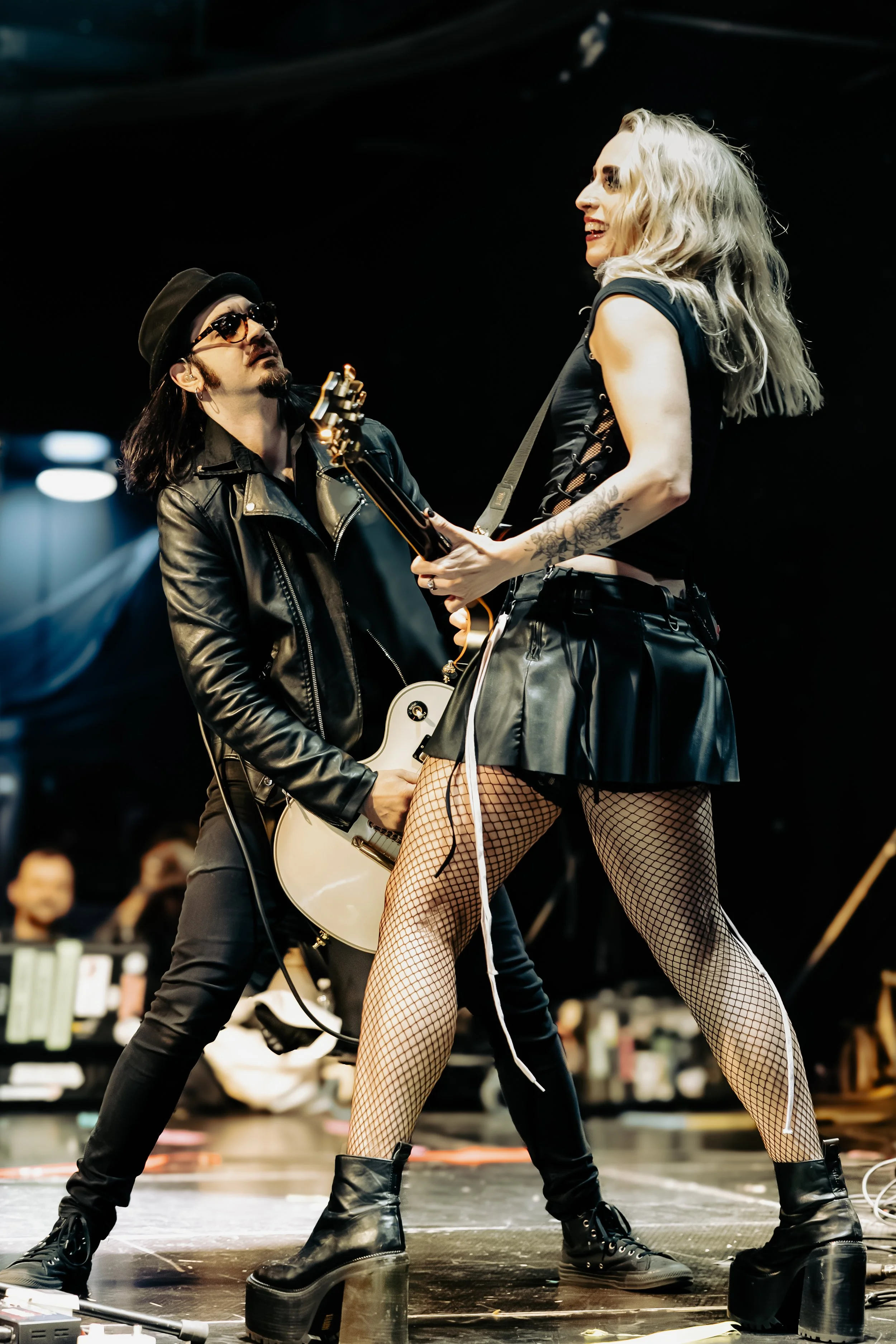 Two musicians playing guitars on stage, one man and one woman, with audience in background.