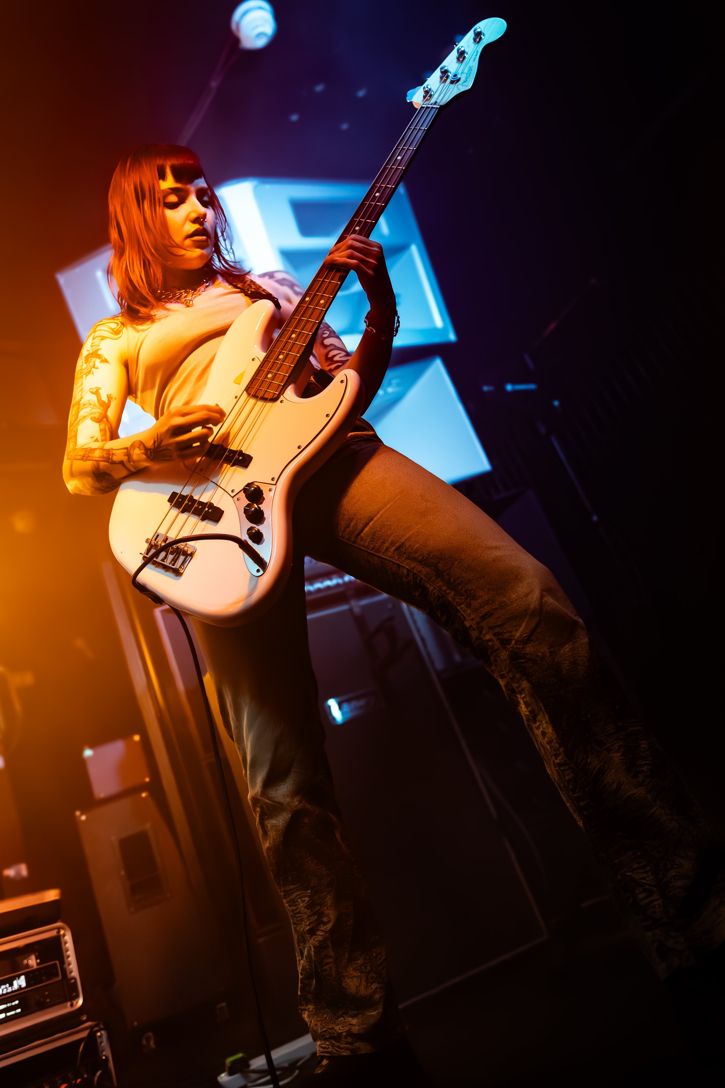 A young woman with tattoos playing a white electric bass guitar on a stage with colorful lighting.
