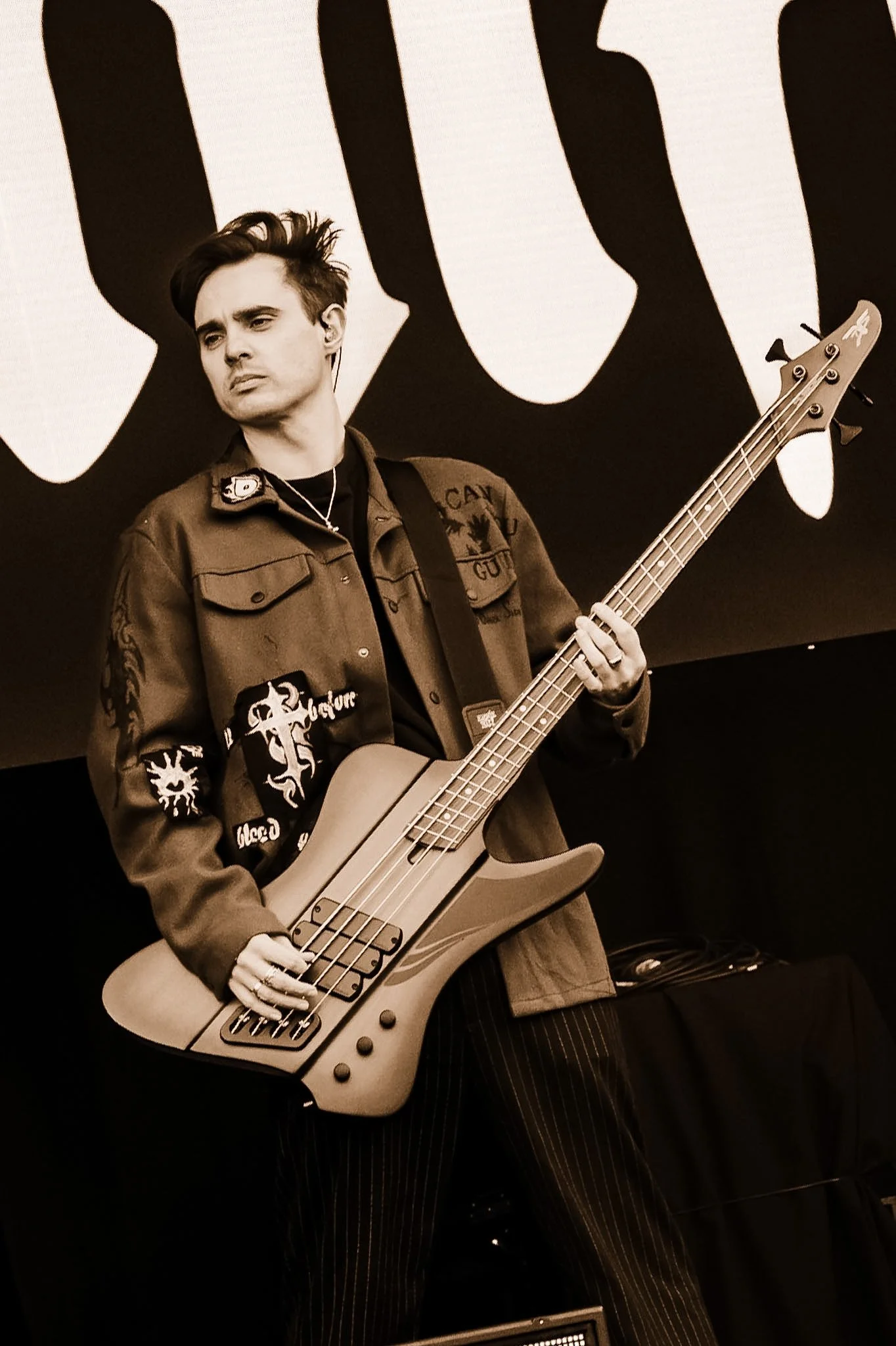 A young man with styled hair playing an electric bass guitar in front of a large backdrop with abstract shapes, wearing a jacket and striped pants.