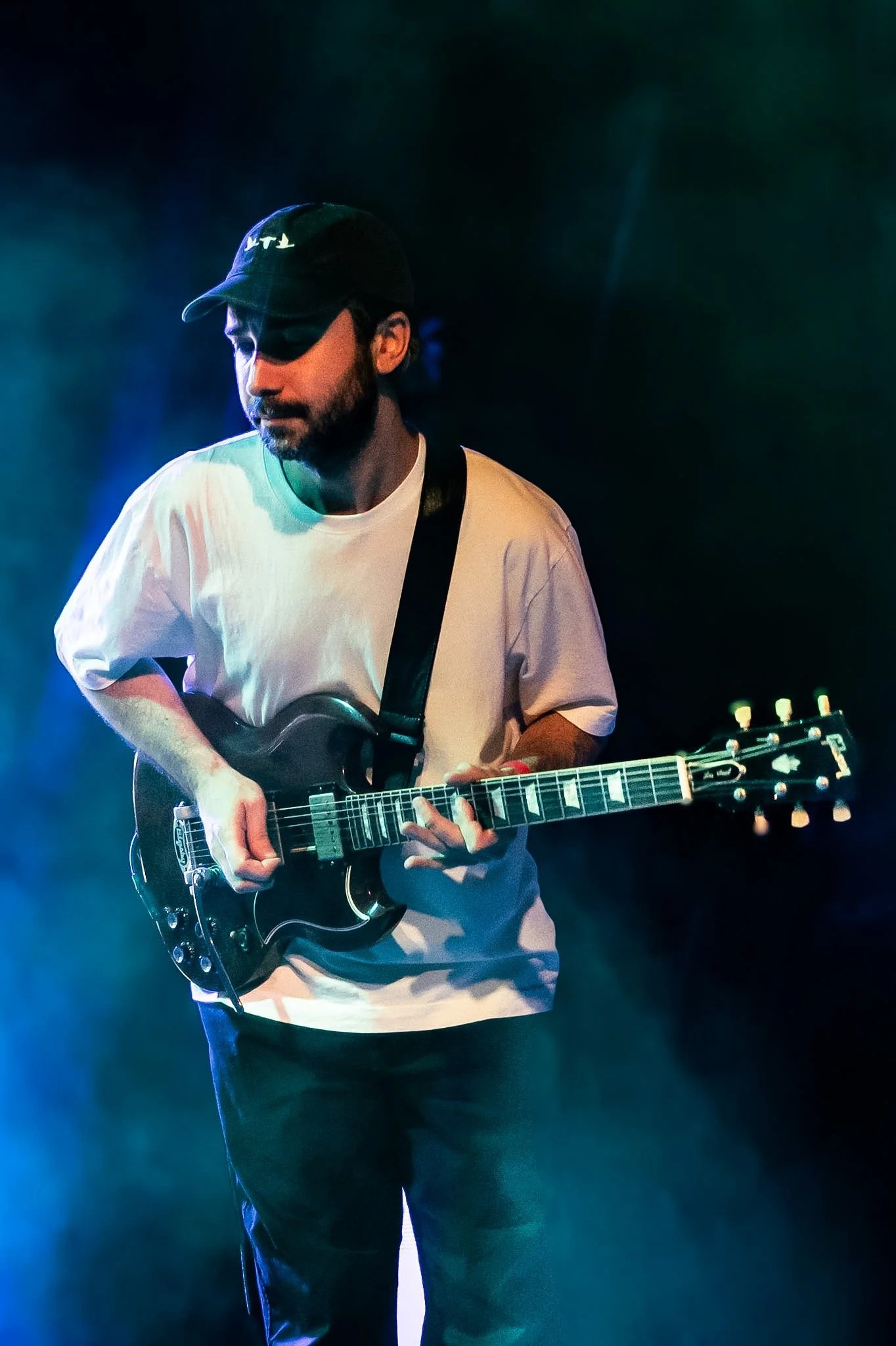 A man wearing a black cap and white T-shirt playing an electric guitar on stage with colorful stage lighting.