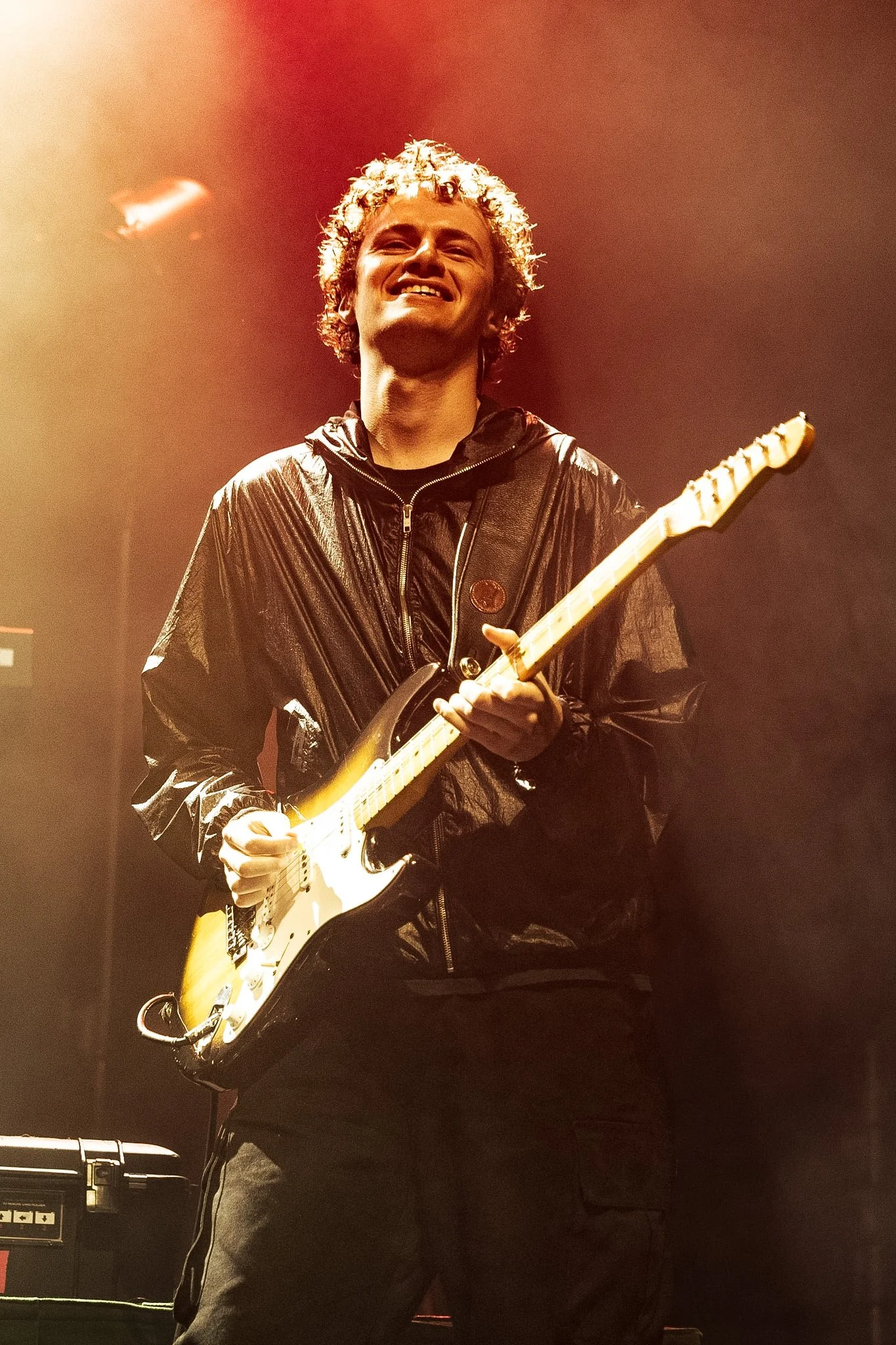 Musician playing an electric guitar on stage, smiling with curly hair, wearing a black shiny jacket. Moio in concert at Koko Camden.