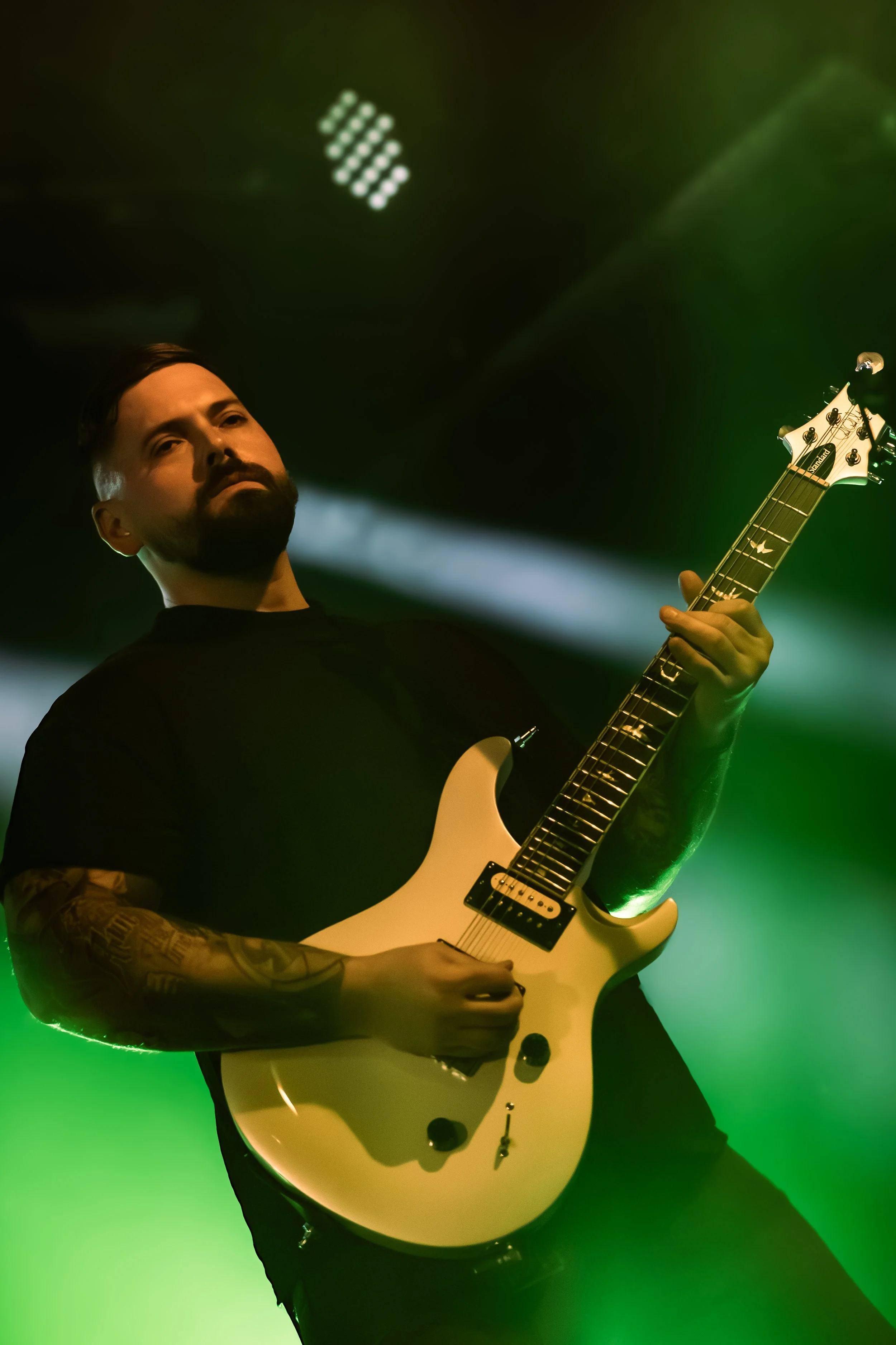 A man with a beard playing an electric guitar on stage with green lighting, wearing a black shirt, with tattoos on his arm, against a dark background with stage lights.