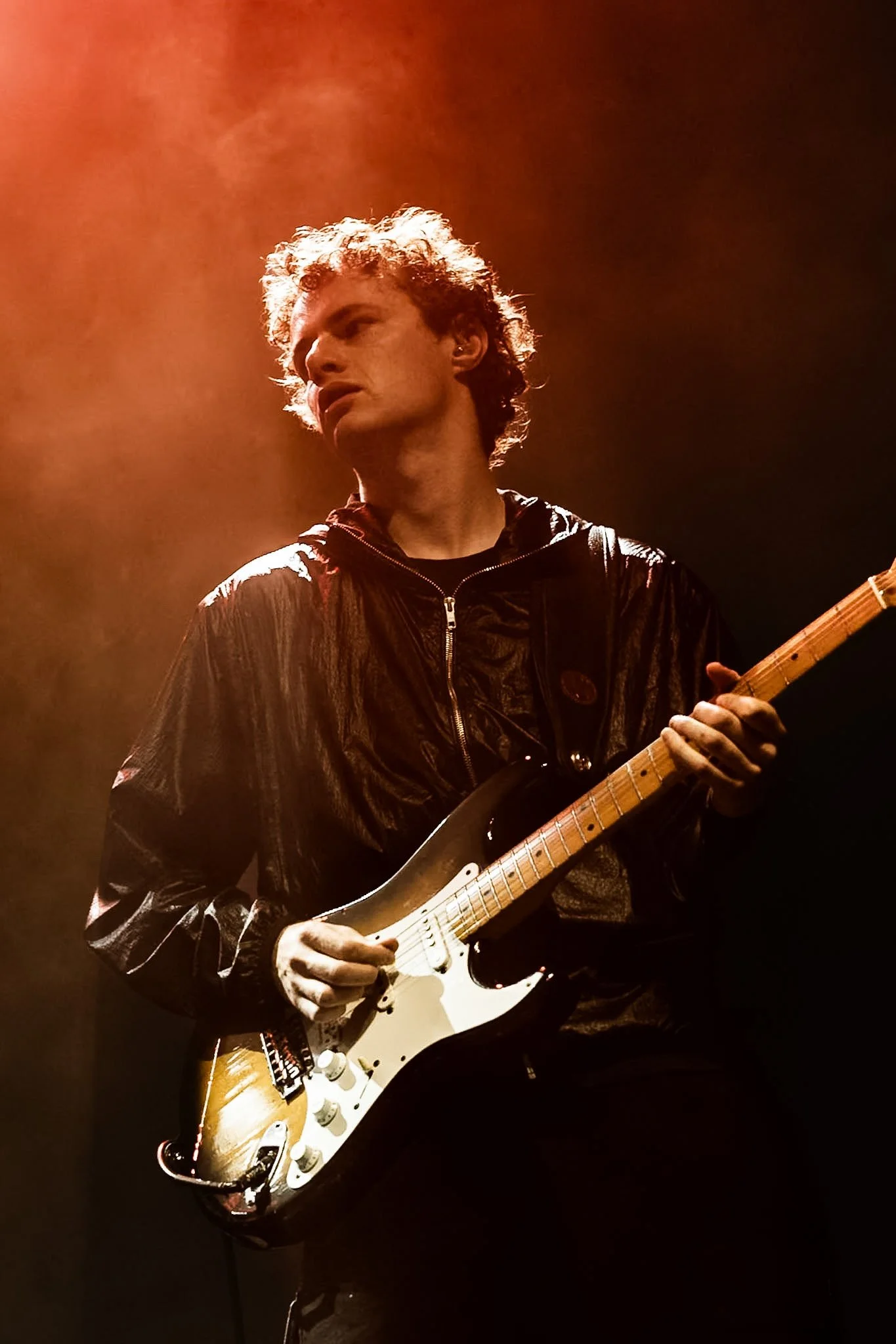 A male musician with curly hair playing an electric guitar on stage, wearing a black jacket with a shiny texture, illuminated by warm stage lighting.