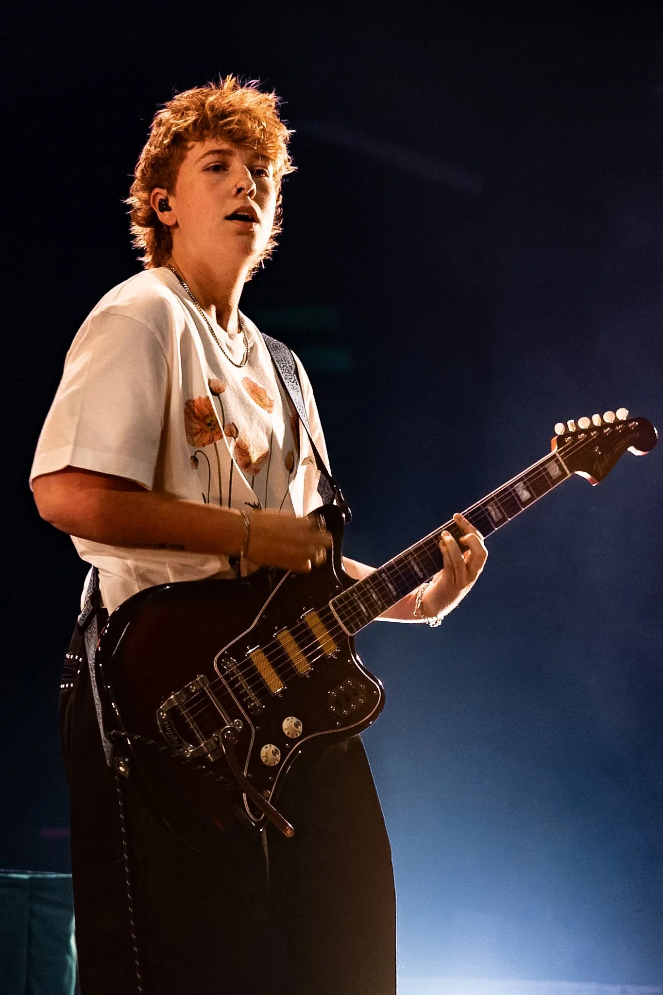 A young person with short, curly red hair playing an electric guitar. They are wearing a white t-shirt with a floral pattern and a silver chain necklace, and they have in-ear monitors. The background is dark with a hint of the stage lighting.