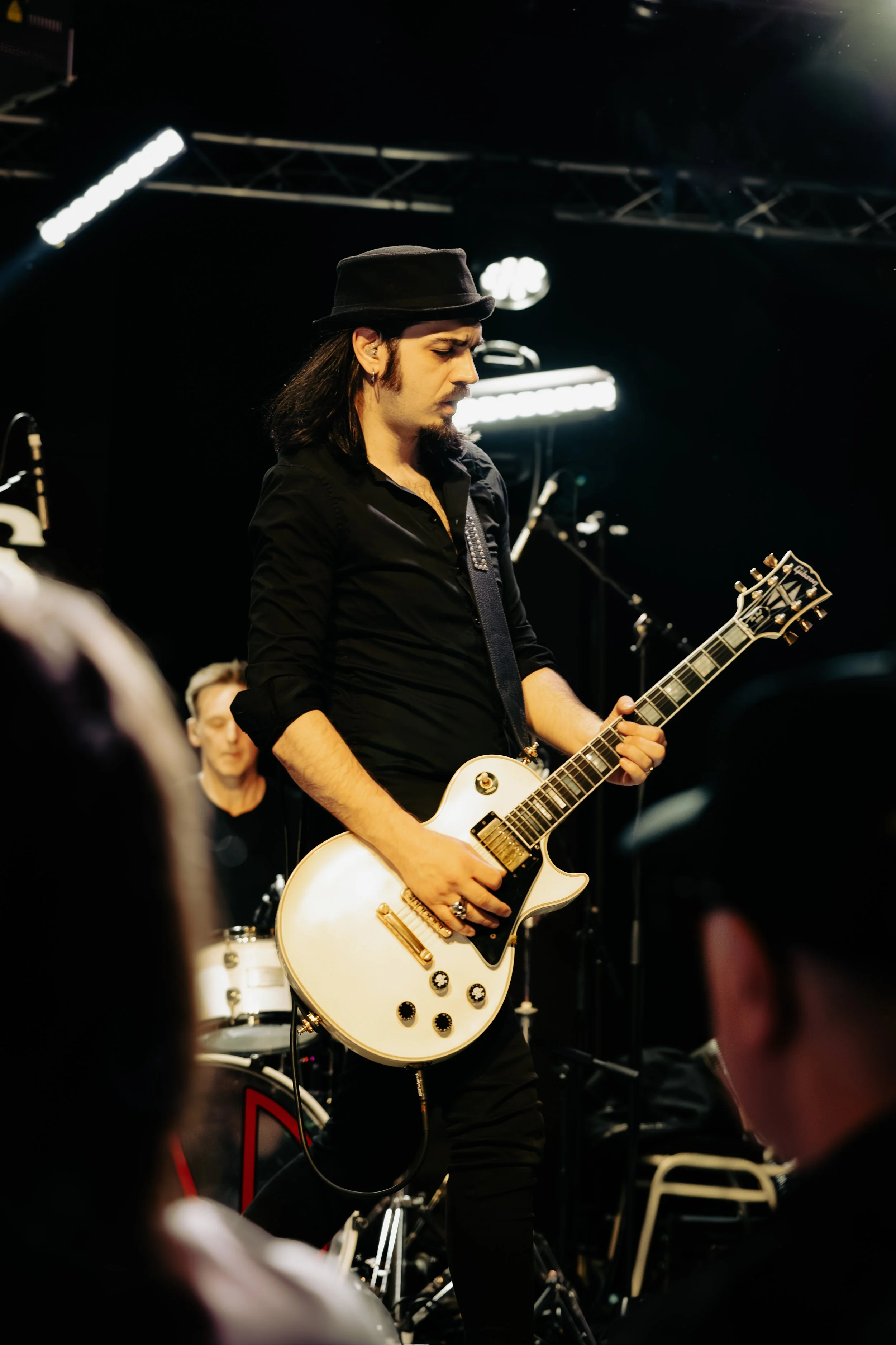 A male guitarist with long hair, a black hat, and earrings, playing a white electric guitar during a concert, with stage lights in the background.