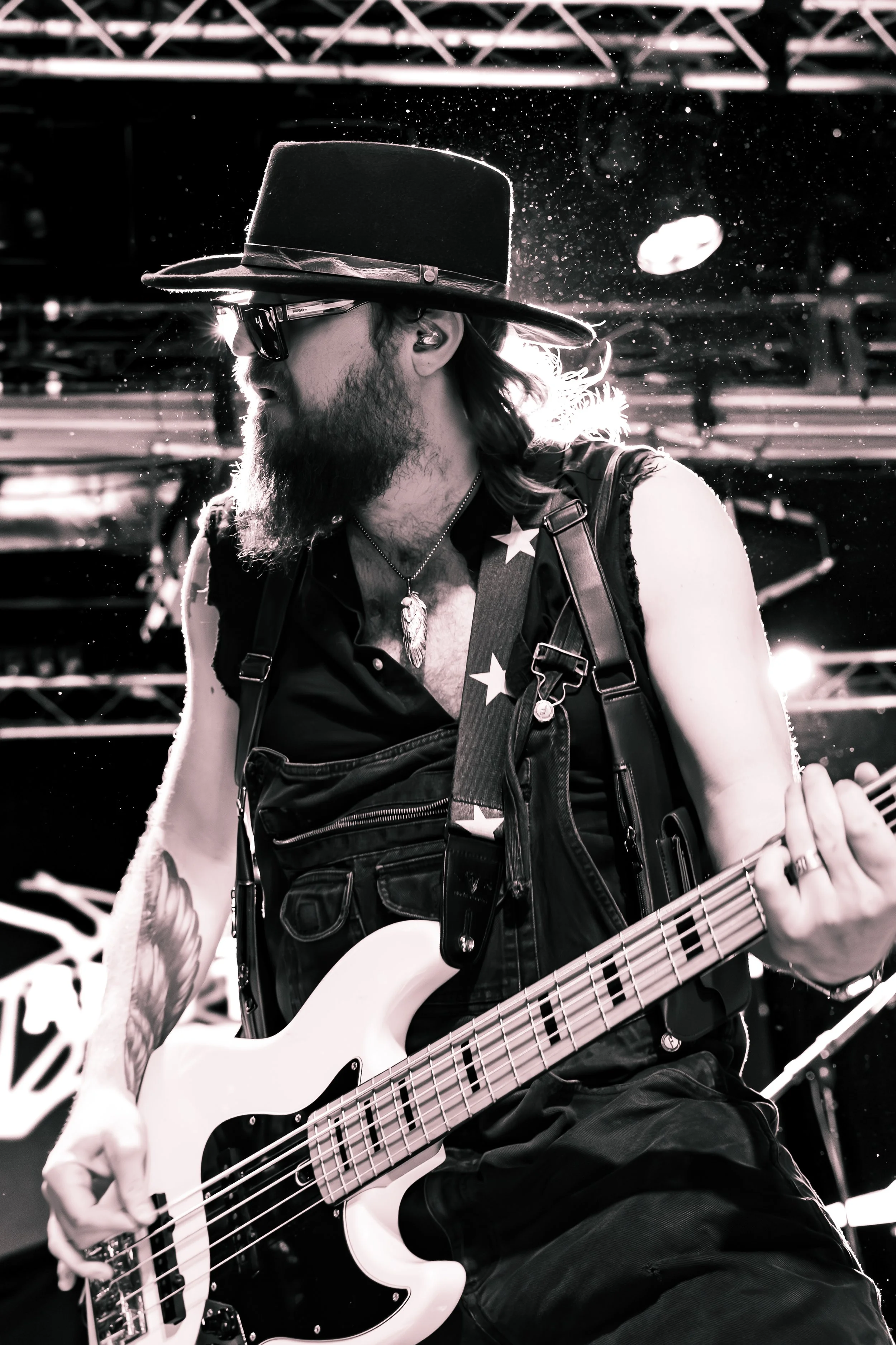 A man with long hair, a beard, wearing sunglasses, a wide-brimmed hat, a sleeveless vest, and a star-patterned bandana playing an electric bass guitar on stage, with stage lights and equipment in the background.