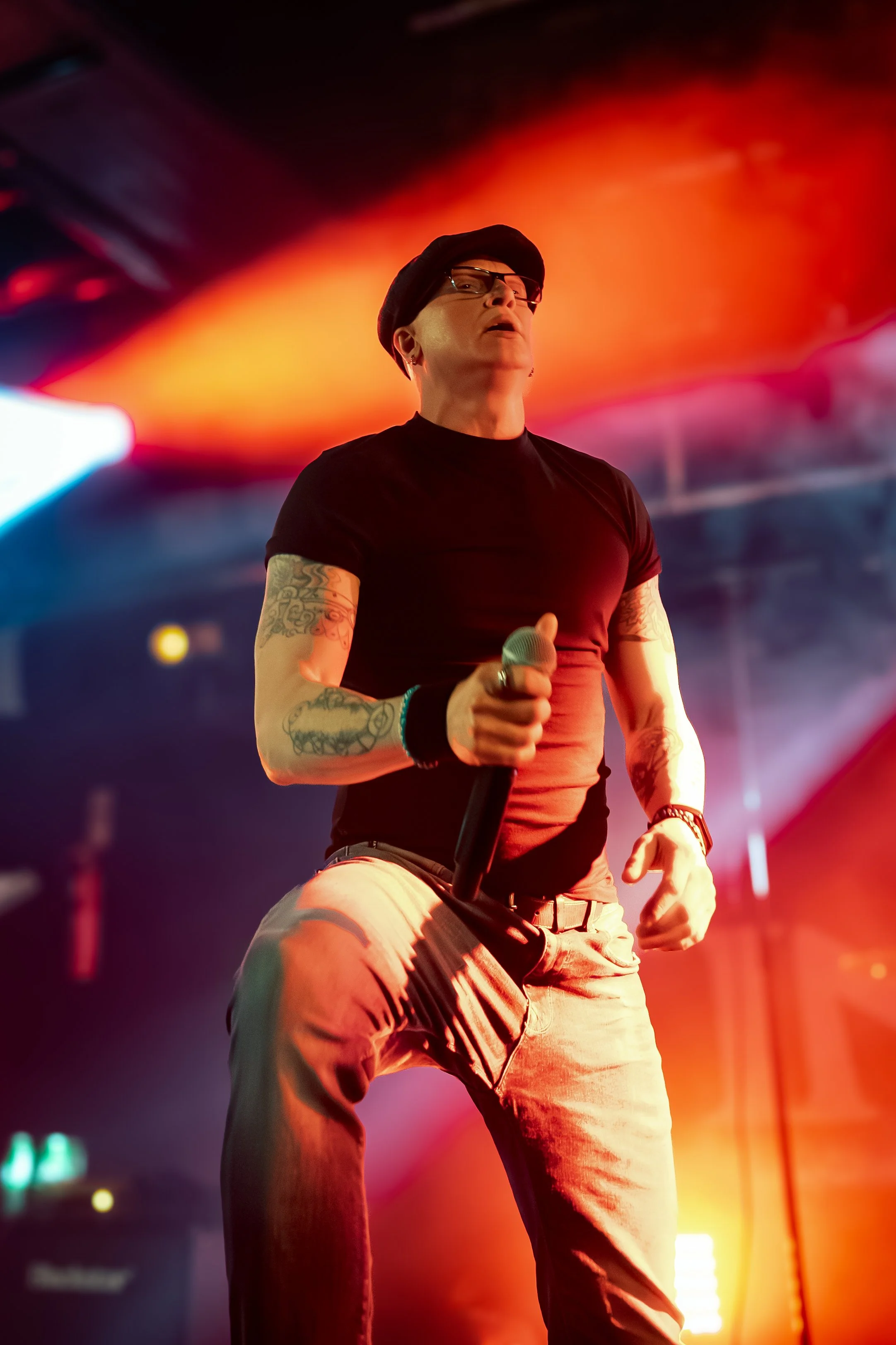 A person holding a microphone on stage with colorful red, orange, and purple lighting in the background.