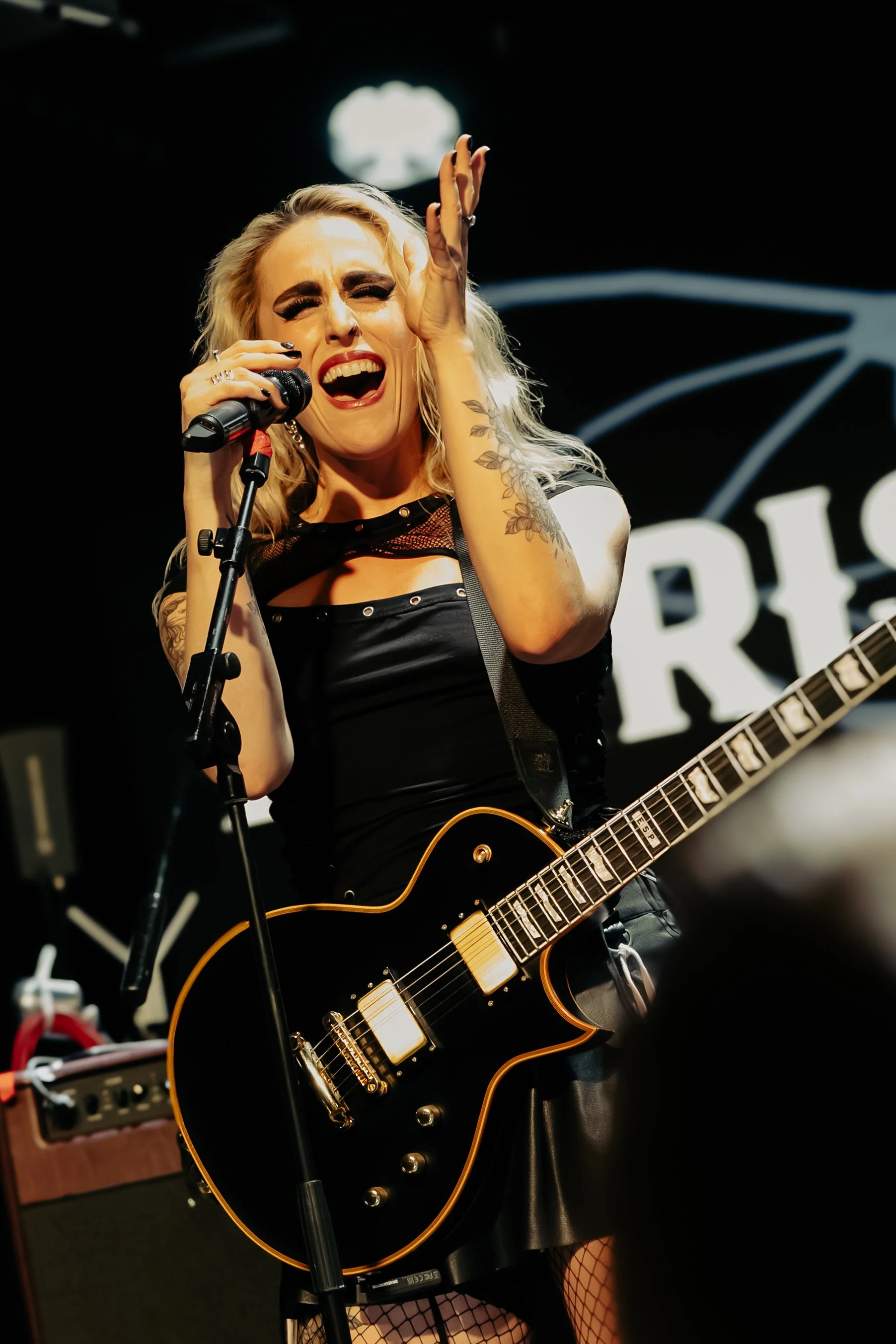 A woman with blonde hair and tattoos singing passionately into a microphone while playing a black and gold electric guitar on stage.
