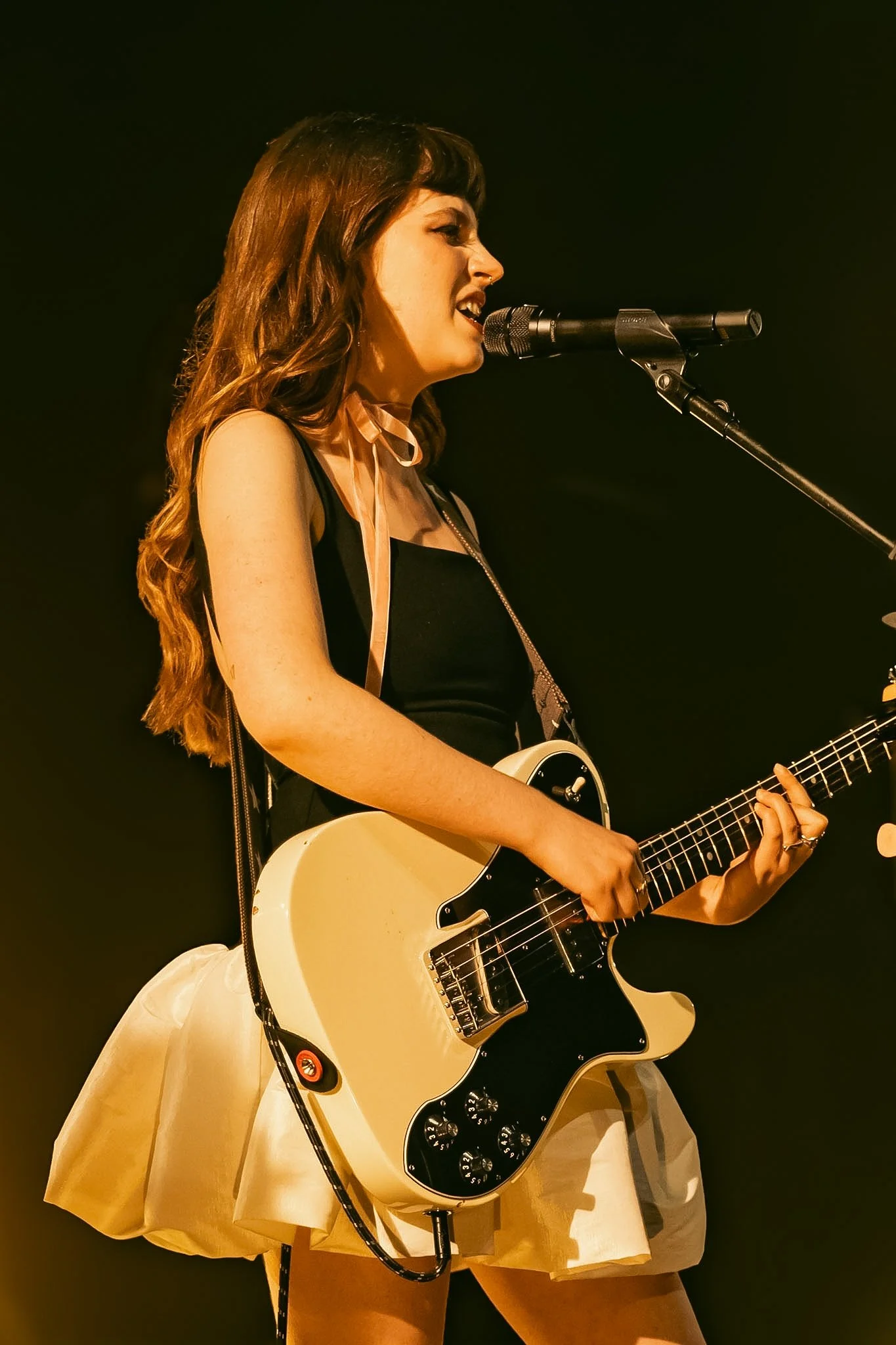 A female musician singing into a microphone while playing an electric guitar on stage.