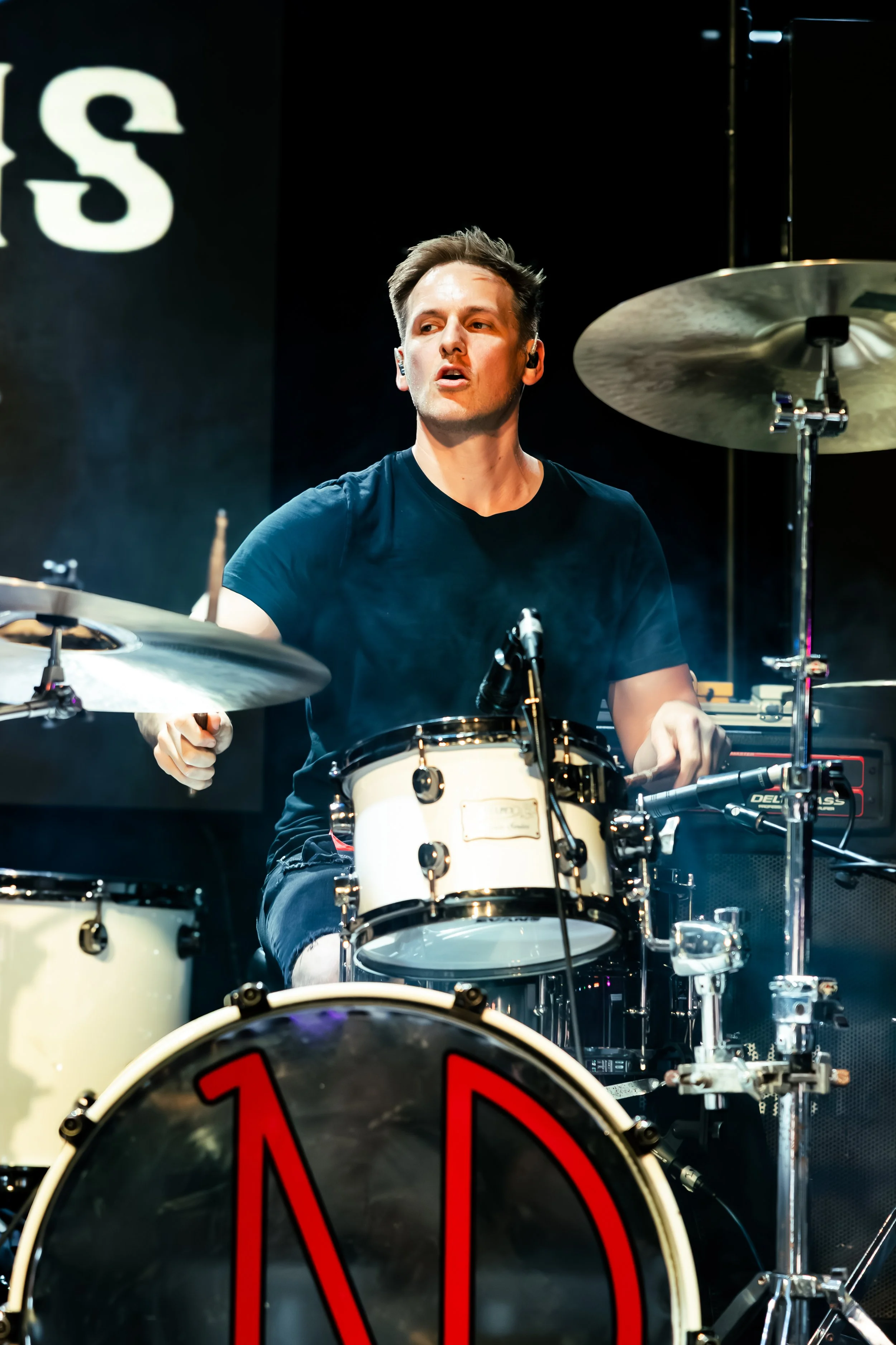 A young man is playing drums on stage, wearing a black t-shirt, with a focused expression, surrounded by various drum equipment, with a dark backdrop and stage lighting.