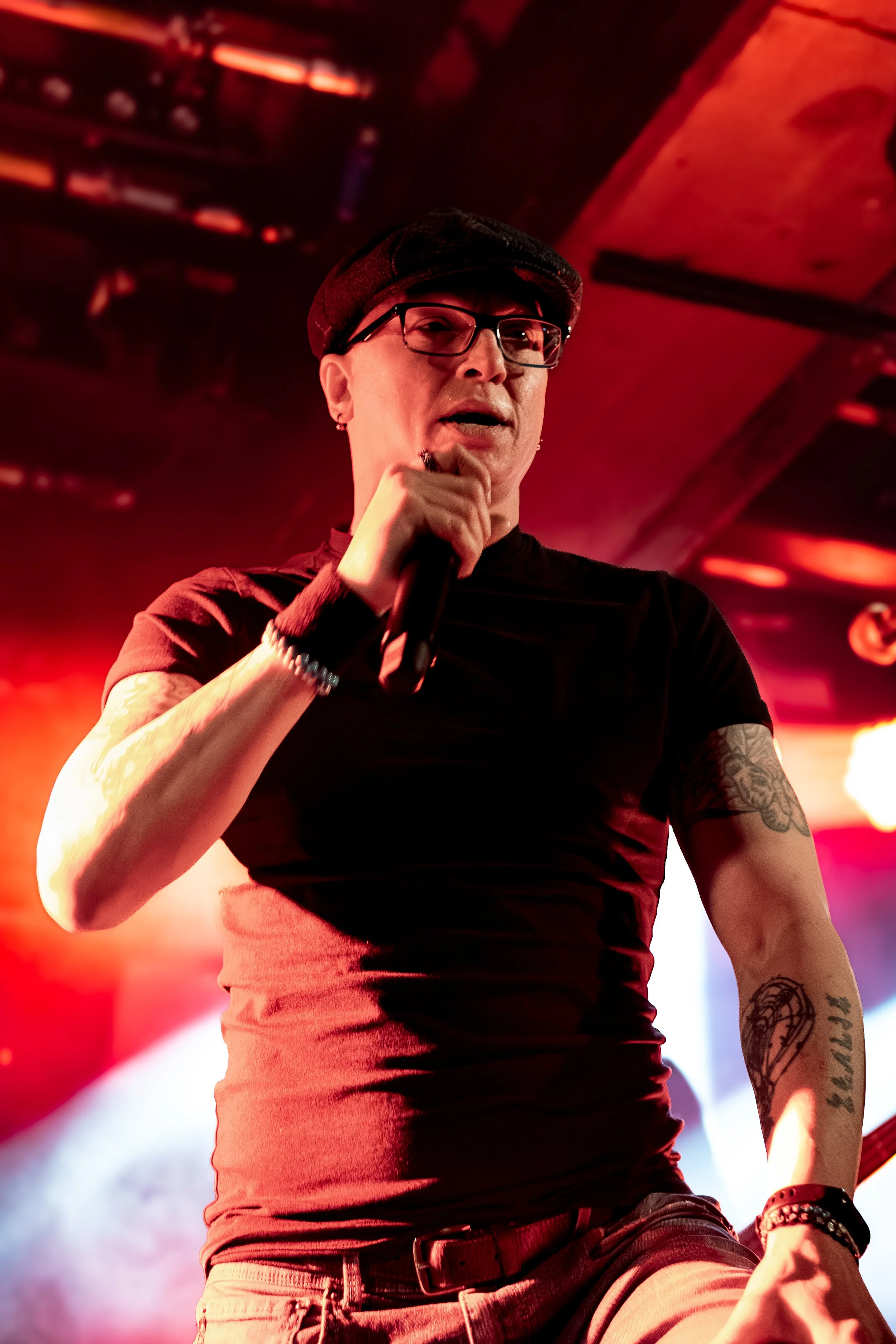 A person with tattoos on their arm, wearing glasses, a cap, and a black T-shirt, holding a microphone, appears to be performing on stage with colorful stage lighting.